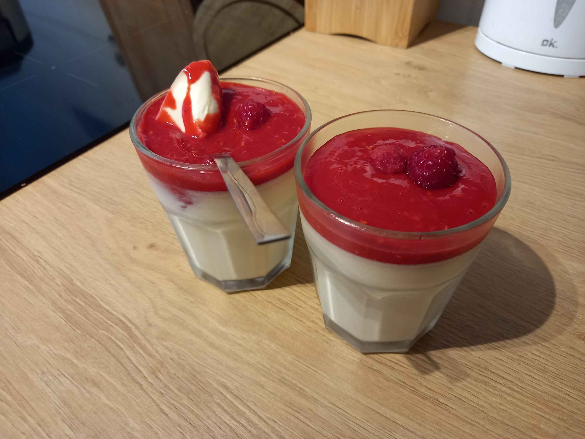 An image of 2 glasses full of white panna cotta, topped with red sauce made from raspberries. The dish is topped with some whole raspberries for decoration, and the glasses stand on a wooden kitchen work surface. 
One of the desserts has a spoon in it, with a dollop of panna cotta on it, arranged to show off the firm consistency.