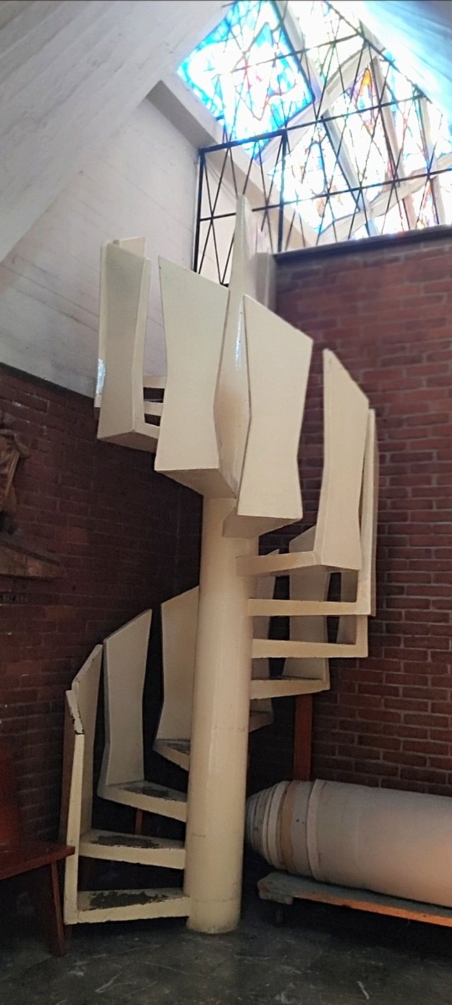 Cream colored freestanding spiral staircase leading up to a higher floor,.lit with stained glass; the banister panels lowok vaguely boney