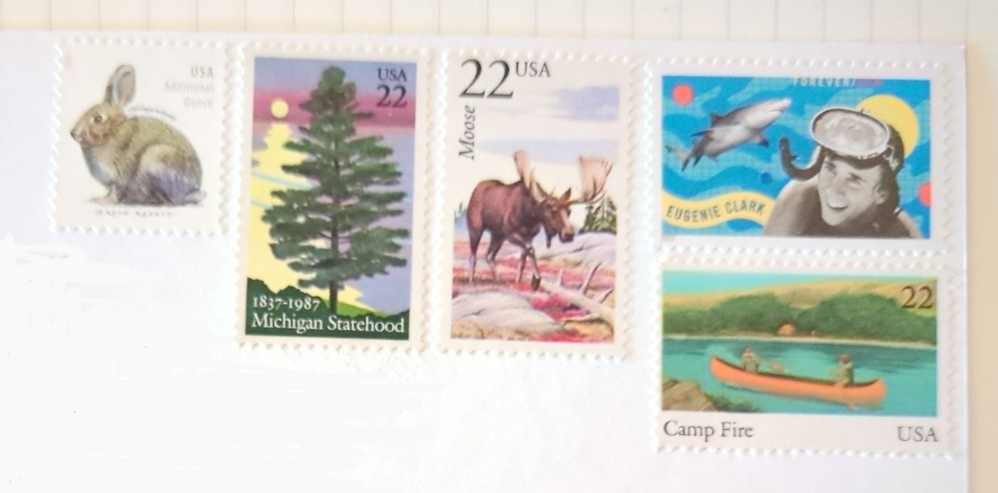 Stamps on the postcard: Eugenie Clark Forever stamp; three 22 cent stamps with a canoe, moose and pine tree; and an additional ounce brush rabbit stamp