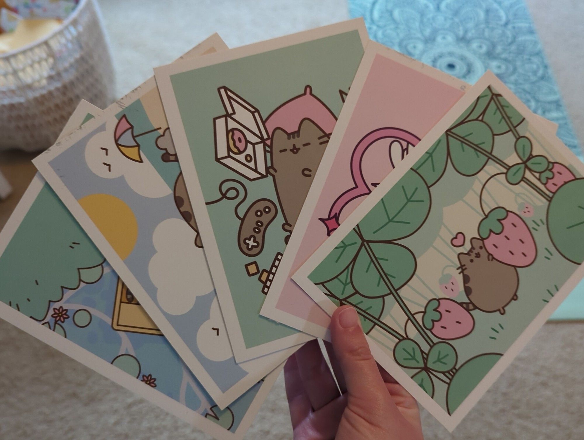 my hand holding 5 Pusheen postcards with pastel green, pink, and blue colors. the top one shows a tiny Pusheen in a forest of strawberry plants. the middle one shows Pusheen fell asleep while gaming and eating donuts.