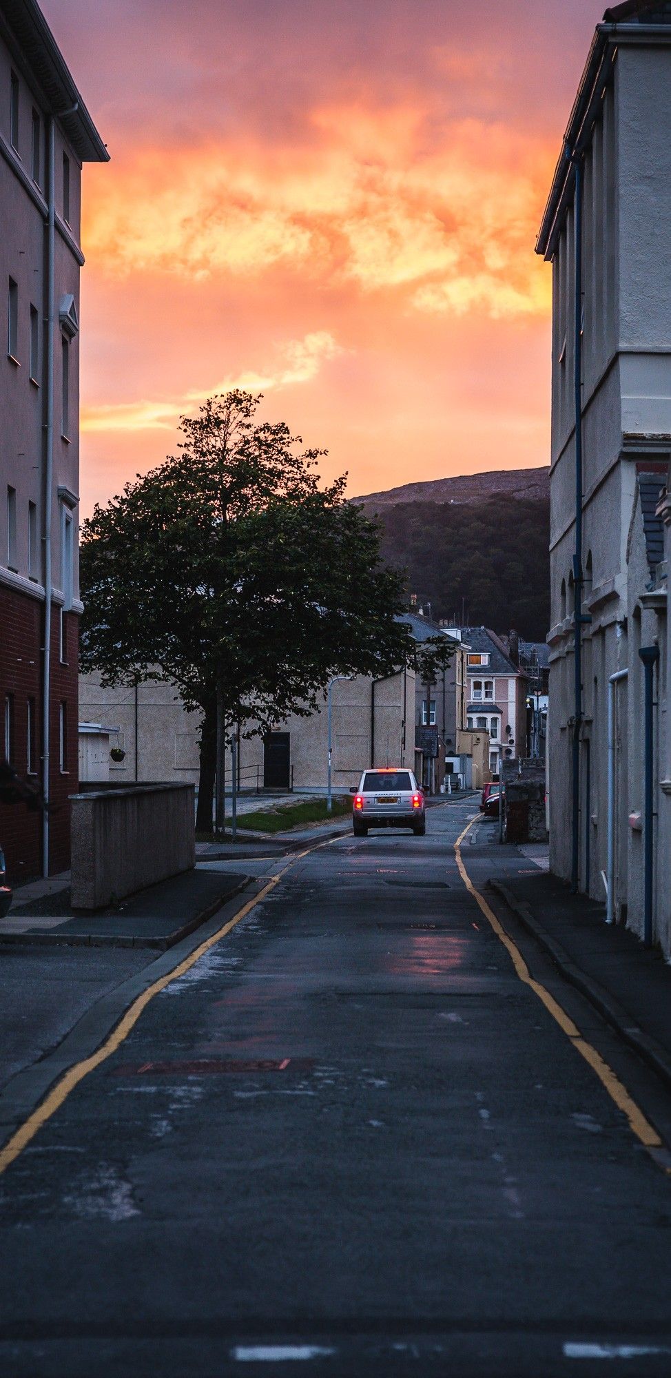 A narrow road passing between two buildings, the red brake lights of a Range Rover, the sky lit up with the setting sun.