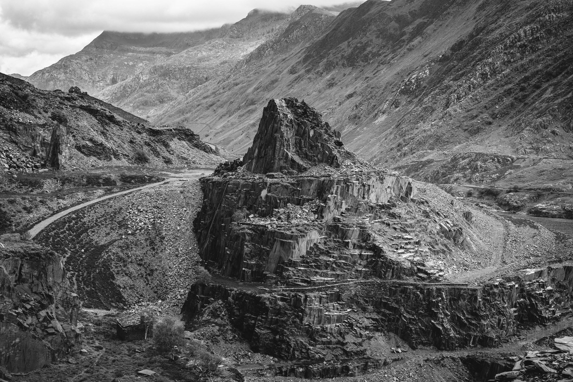 A black and white landscape photo. A scarred and brutal landscape, the results of industrial slate mining. Terraced tracks circle a slate column that emerges from slate spoil heaps, in the distance the sides of a steep valley in the mountains of Eryri.