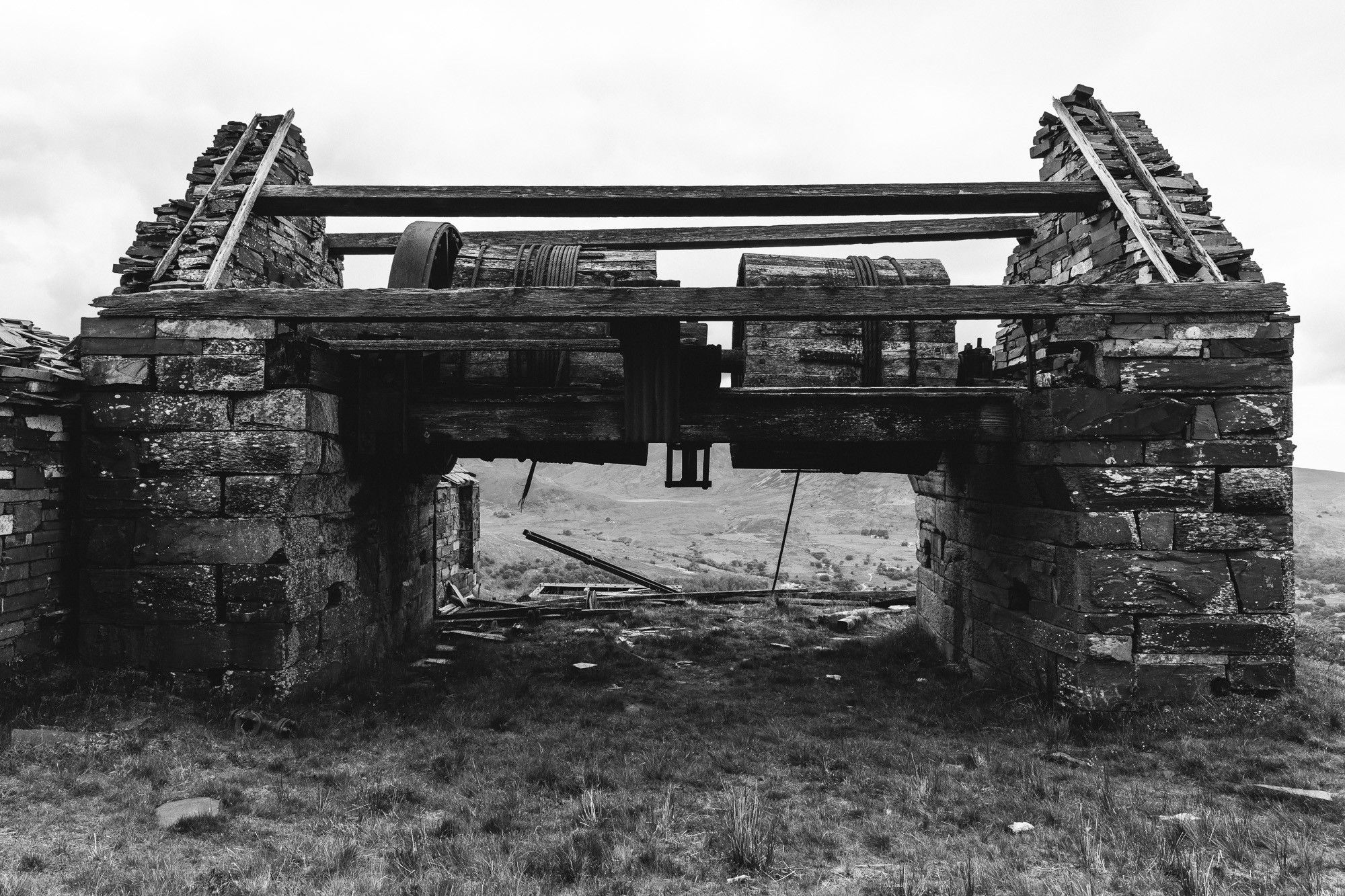 A black and white photo of a derelict winding house high up in the hills of north Wales that once pulled wagons for the slate quarry up and down inclines. The wooden drums with some of the steel cable are still visible between the slate walls.