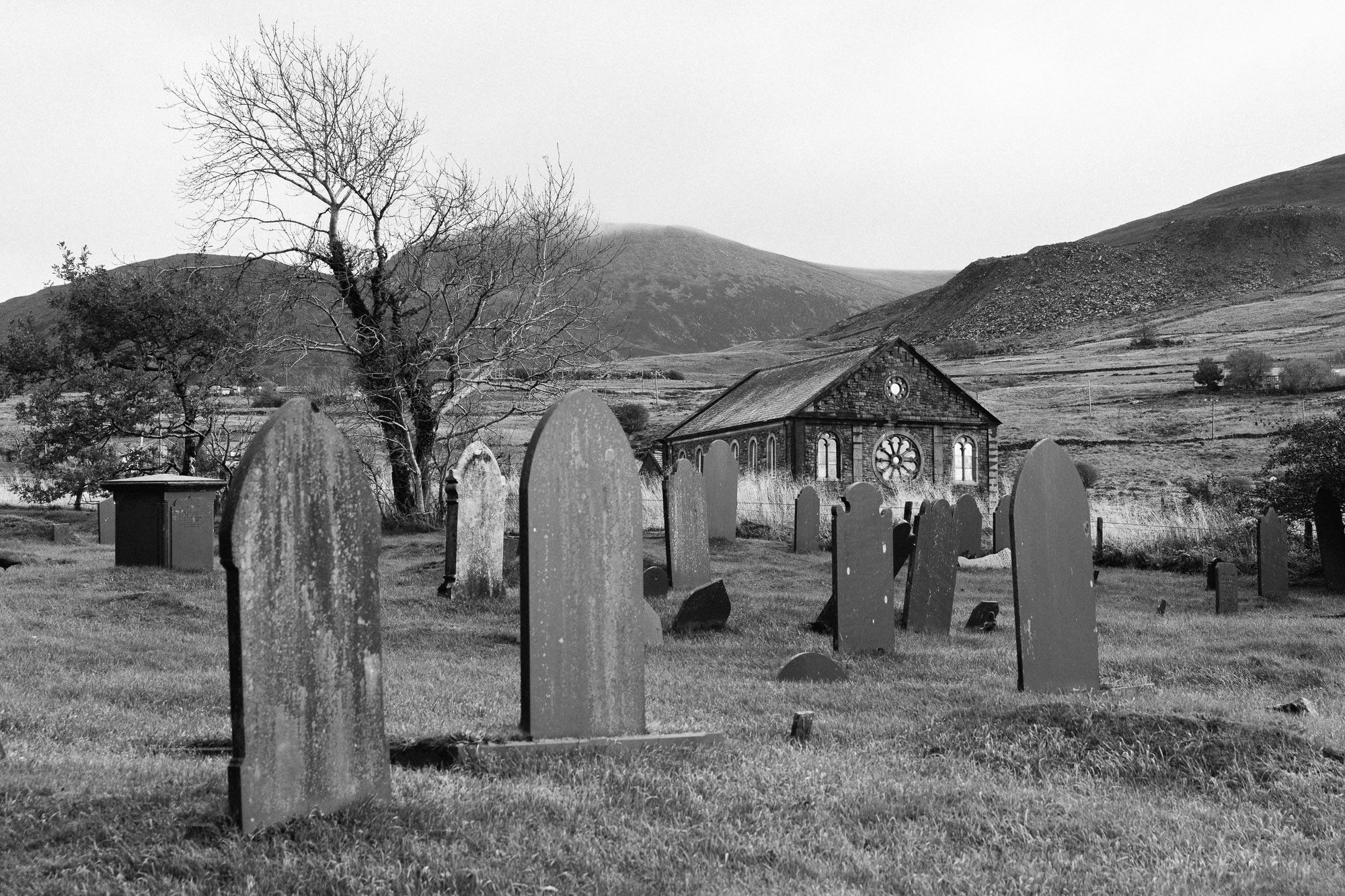 Slate gravestones in a cemetery, in the middle distance a Welsh chapel, in the distance slate tips and mountains.