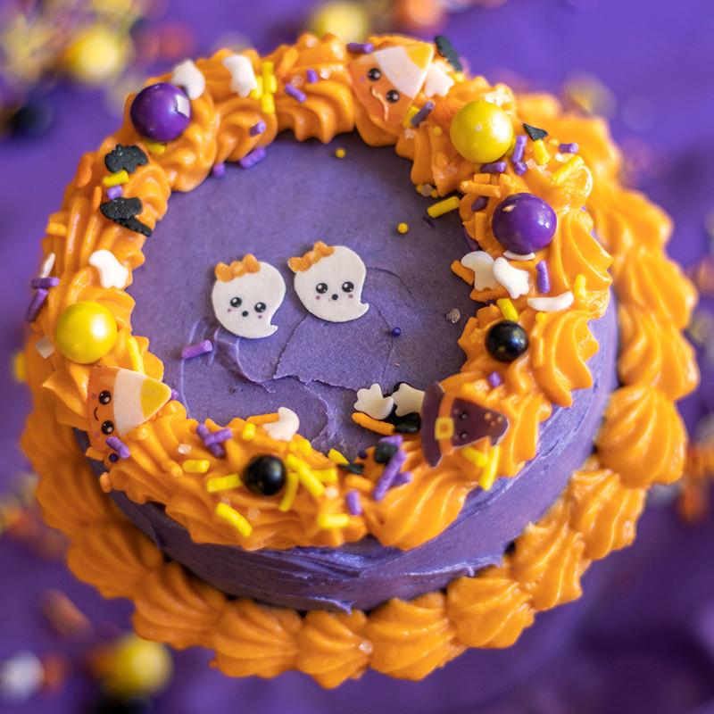 A small Halloween themed cake sits on a purple background. The cake is purple with a bright orange shell border, and has colorful black, yellow, and purple sprinkles. Two little ghosts made of sugar are atop the cake.