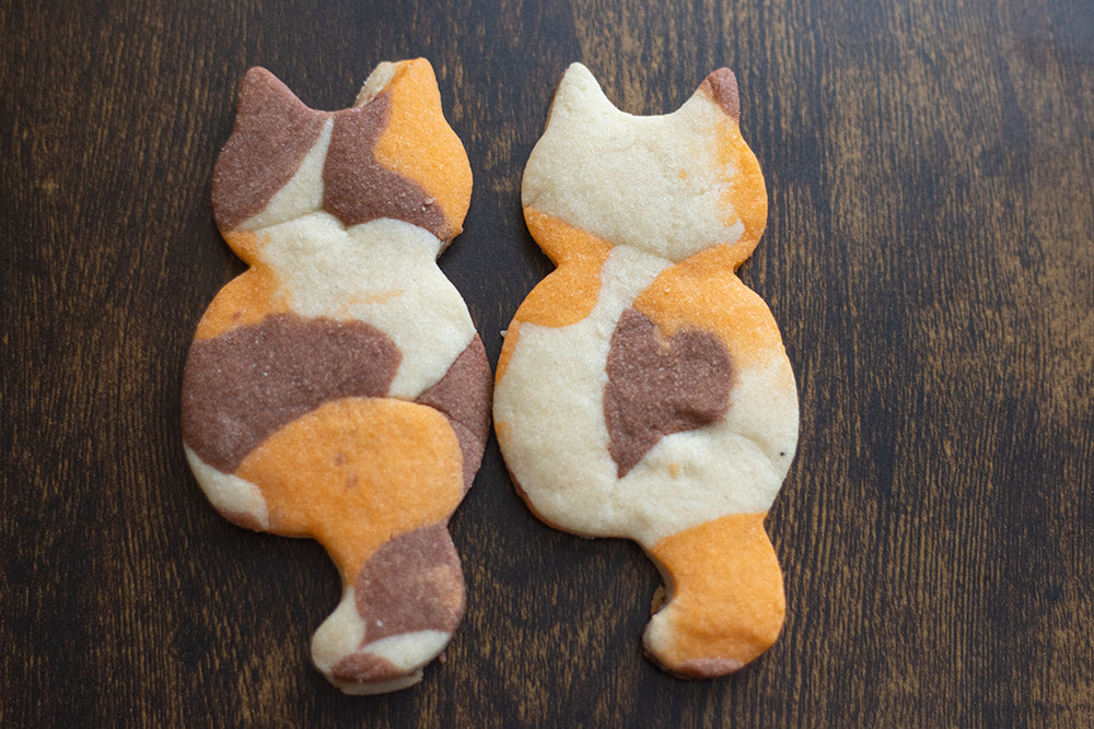 Two cookies shaped like cats sit on a dark wood table. They are marbled colors of brown, orange, and tan. They resemble a calico cat.