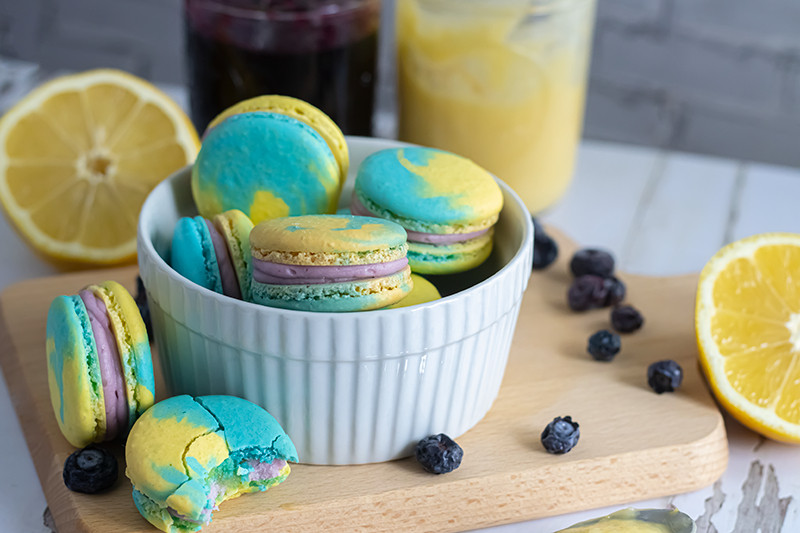 On a weathered white wood plank surface, a light colored wood cutting board contains scattered blueberries and sliced lemons, surrounding a white rammekin filled with macarons. The macarons have light blue and yellow shells, the colors are swirled together. The filling is a light purple. In the background, unfocused, are two jars - one of a yellow lemon curd, and the other a dark purple blueberry jam.