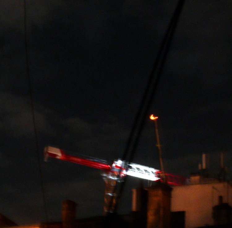 photo de nuit et un peu floue, une grue rouge et blanche pas encore complète avec un grand treuil à côté, les toits des maisons et immeubles environnants en-dessous, un ciel de nuit un peu voilé au-dessus
Une partie de la grue est très éclairée par un spot au sol