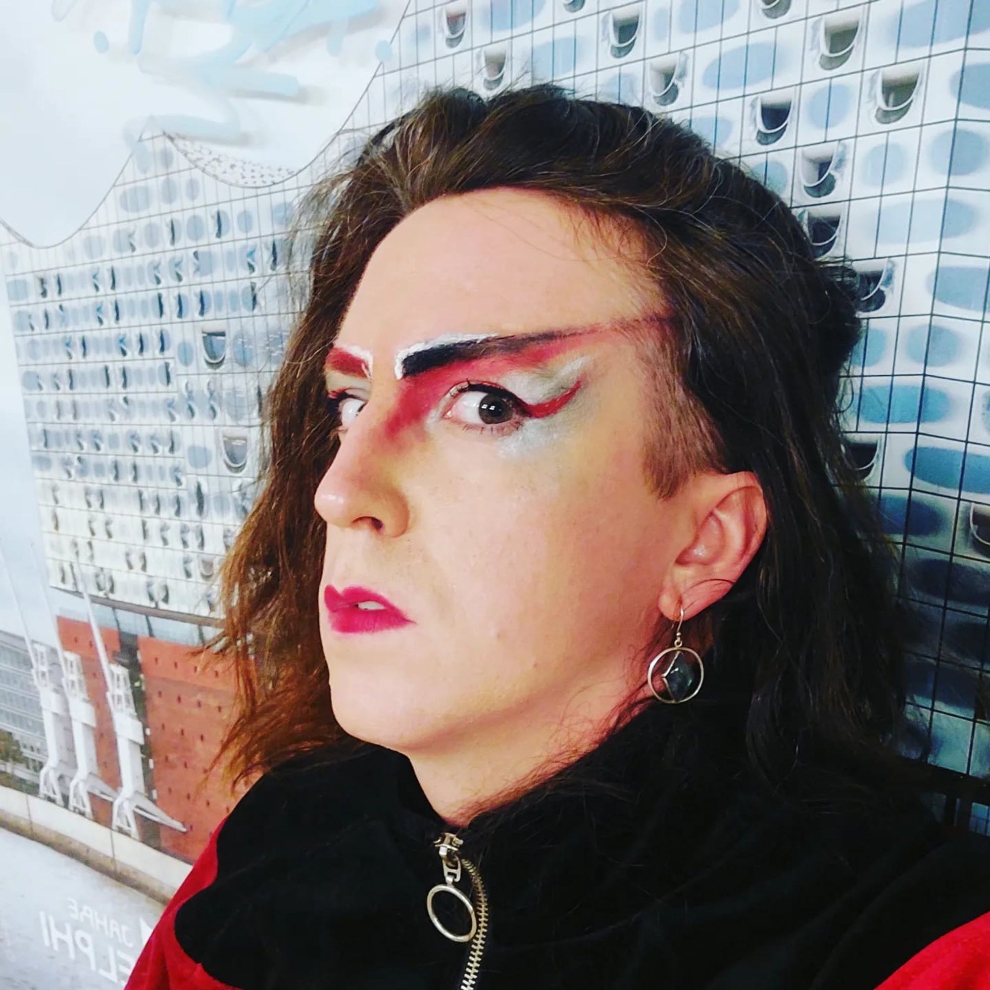 Queer woman with a strong liner makeup with thick black, red and white lines, wearing black and red clothes, in the Berlin underground.