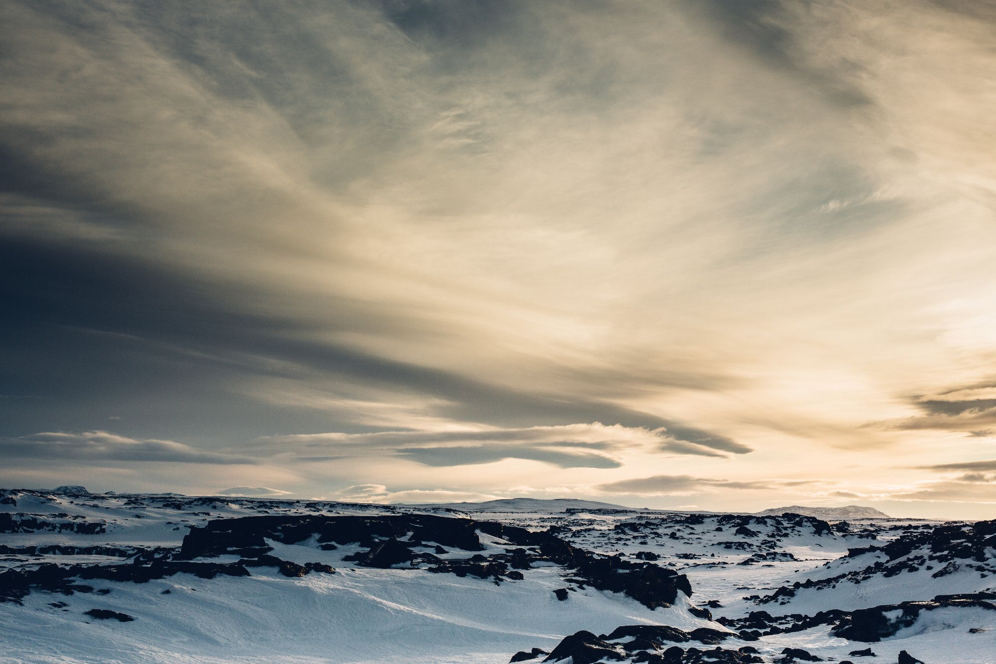 "A dramatic winter landscape featuring snow-covered terrain with dark, rugged rocks scattered across the scene. The sky is filled with dynamic clouds that are illuminated by a soft, golden light, creating a stark contrast between the cold, snowy ground and the warm tones of the sky. The vastness and raw beauty of the environment evoke a sense of isolation and awe." (ChatGPT)