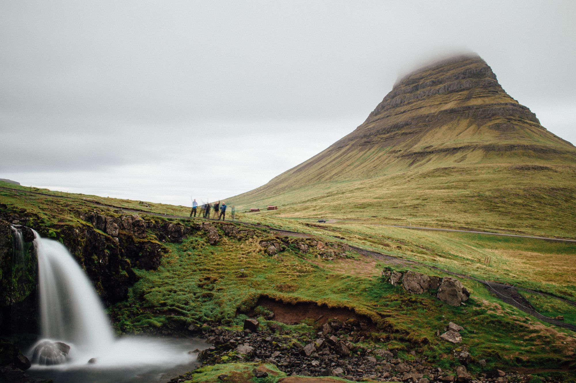 "A misty landscape in Iceland featuring the iconic Kirkjufell mountain, partially obscured by low clouds. In the foreground, a small waterfall cascades into a pool, surrounded by lush green terrain. A group of hikers stands near the edge of the cliff, adding a sense of scale and adventure to the serene scene."