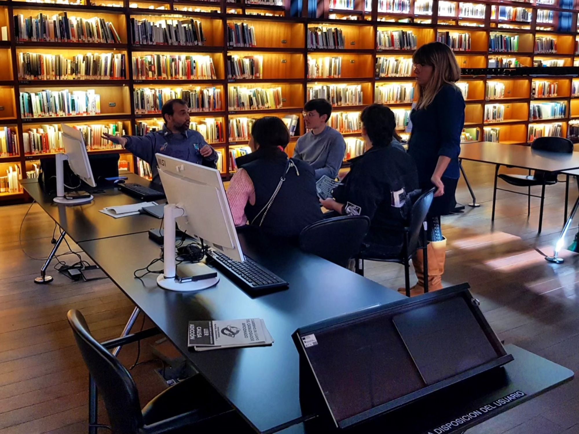 Foto de un grupo de participantes en la biblioteca del Reina Sofía. Se ven dos mesas alargadas y un círculo de personas, de fondo las estanterías retroiluminadas con luz anaranjada de la biblioteca.