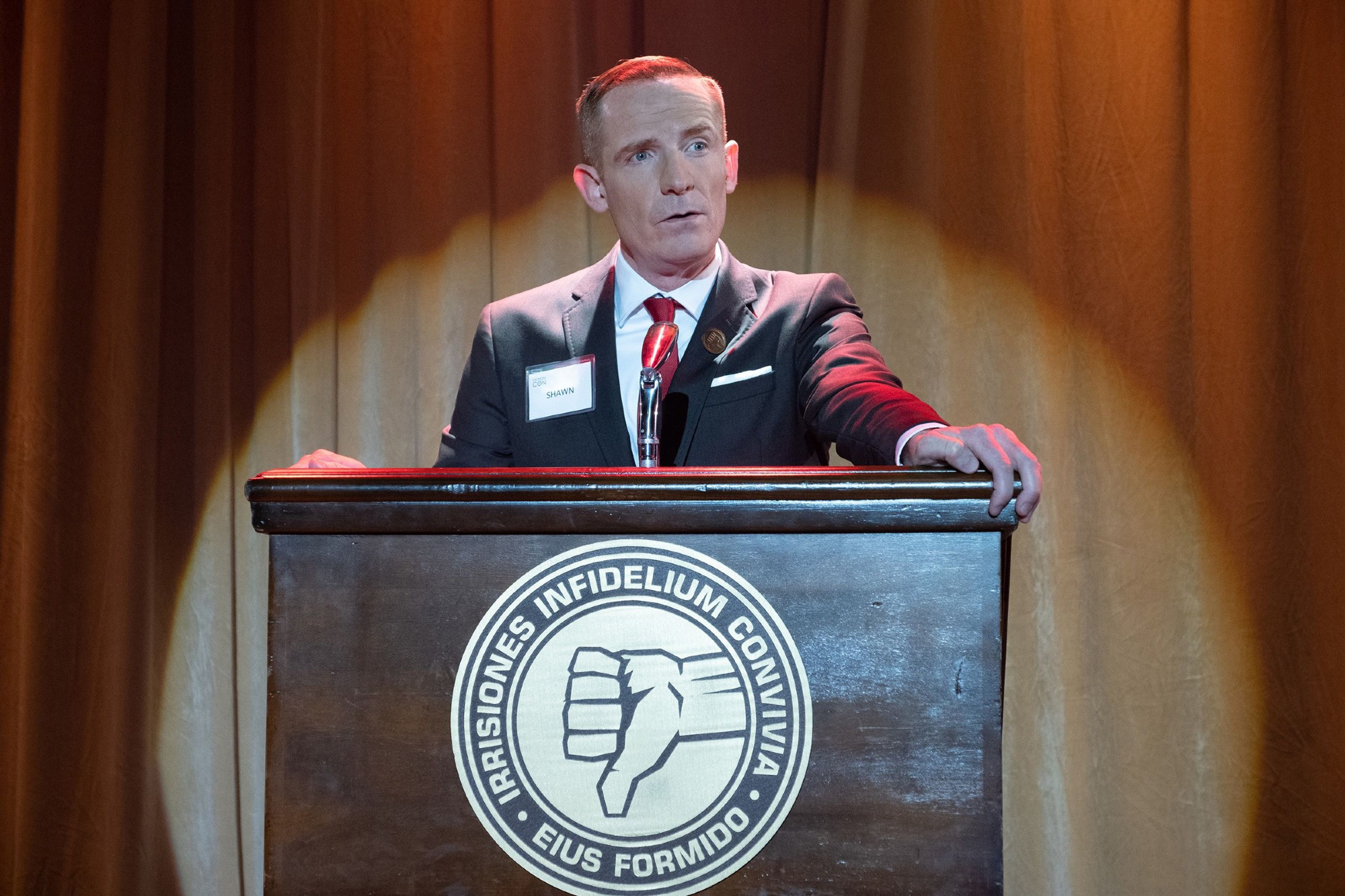 Shawn from The Good Place, at a podium with the thumbs-down symbol and the slogan "irrisiones infidelium convivia - eius formido"