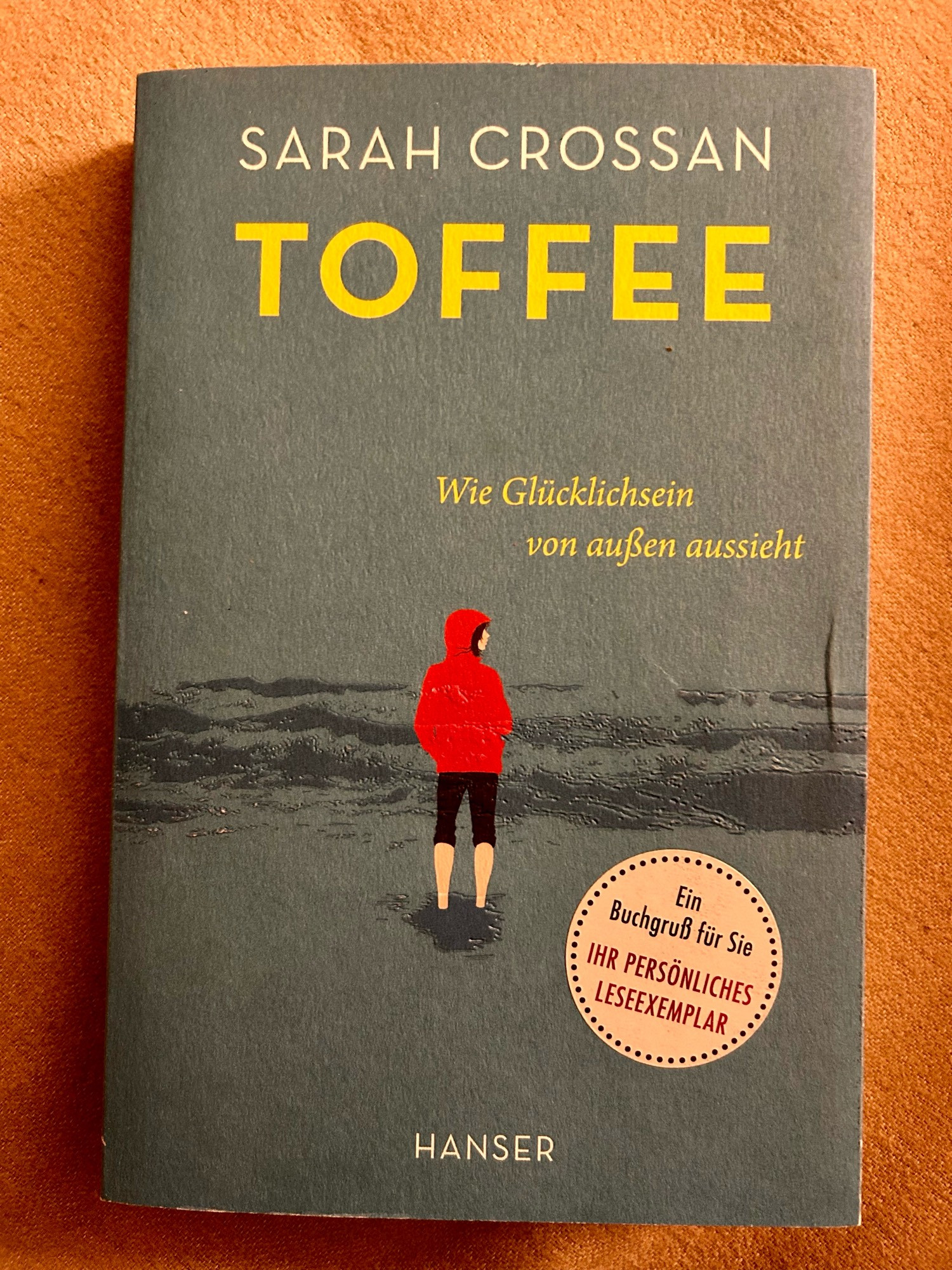 Buchcover von Sarah Crossans „TOFFEE - Wie Glücklichsein von außen aussieht“. Es zeigt ein Mädchen in roter Kapuzenjacke am Strand.