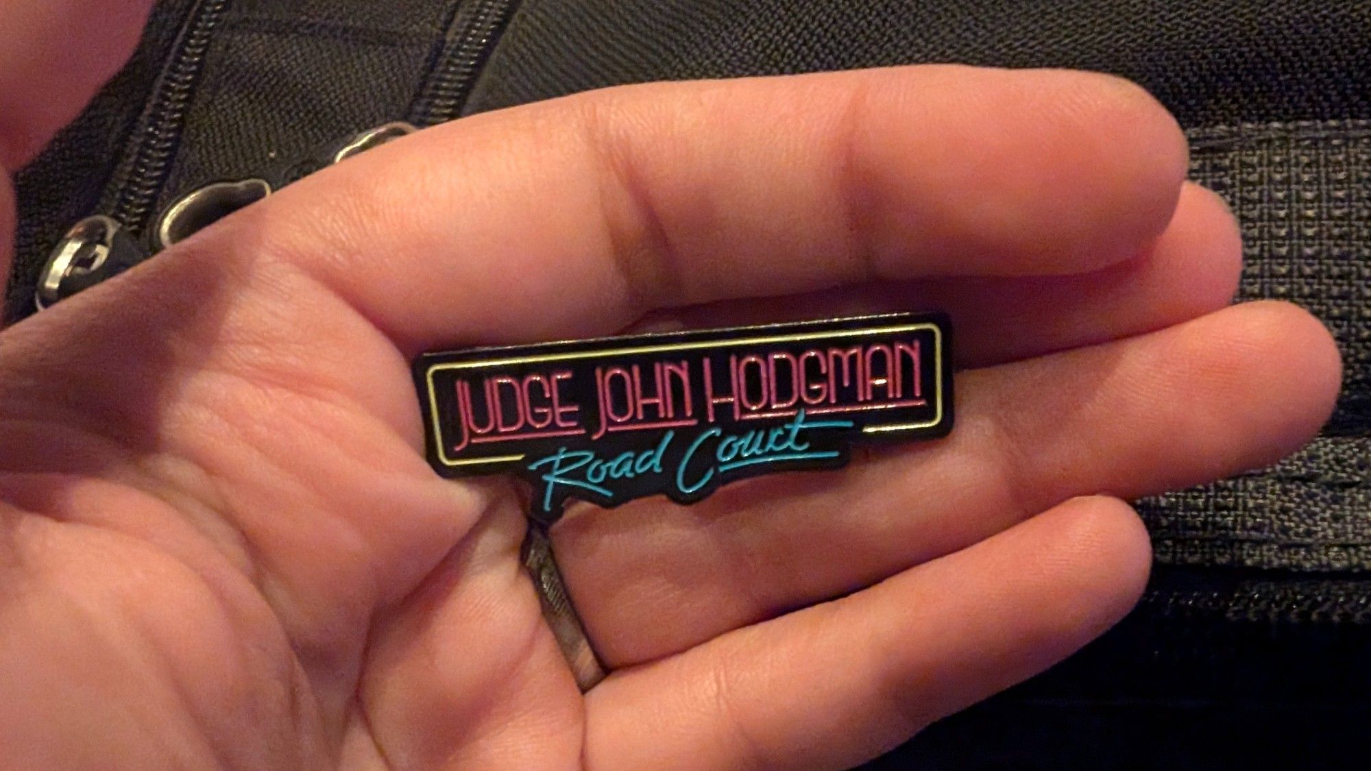 A hand holding a pin that reads Judge John Hodgman Road Court styled like neon lights.