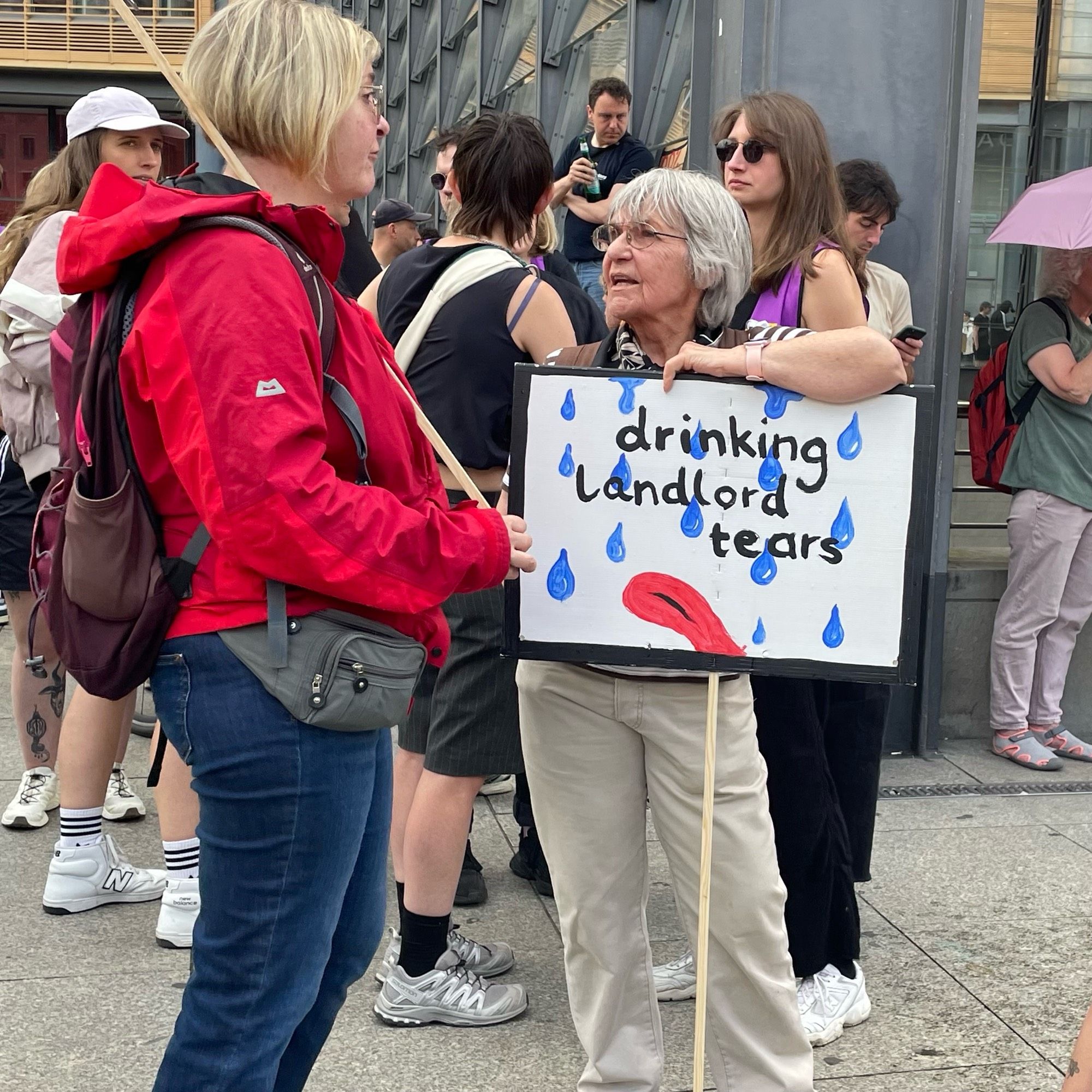 Zwei Frauen sind einander zugewandt. Eine lehnt auf einem Plakat an einer Stange auf dem blaue Tropfen und unten eine rote Zunge gemalt sind „drinking landlord tears“