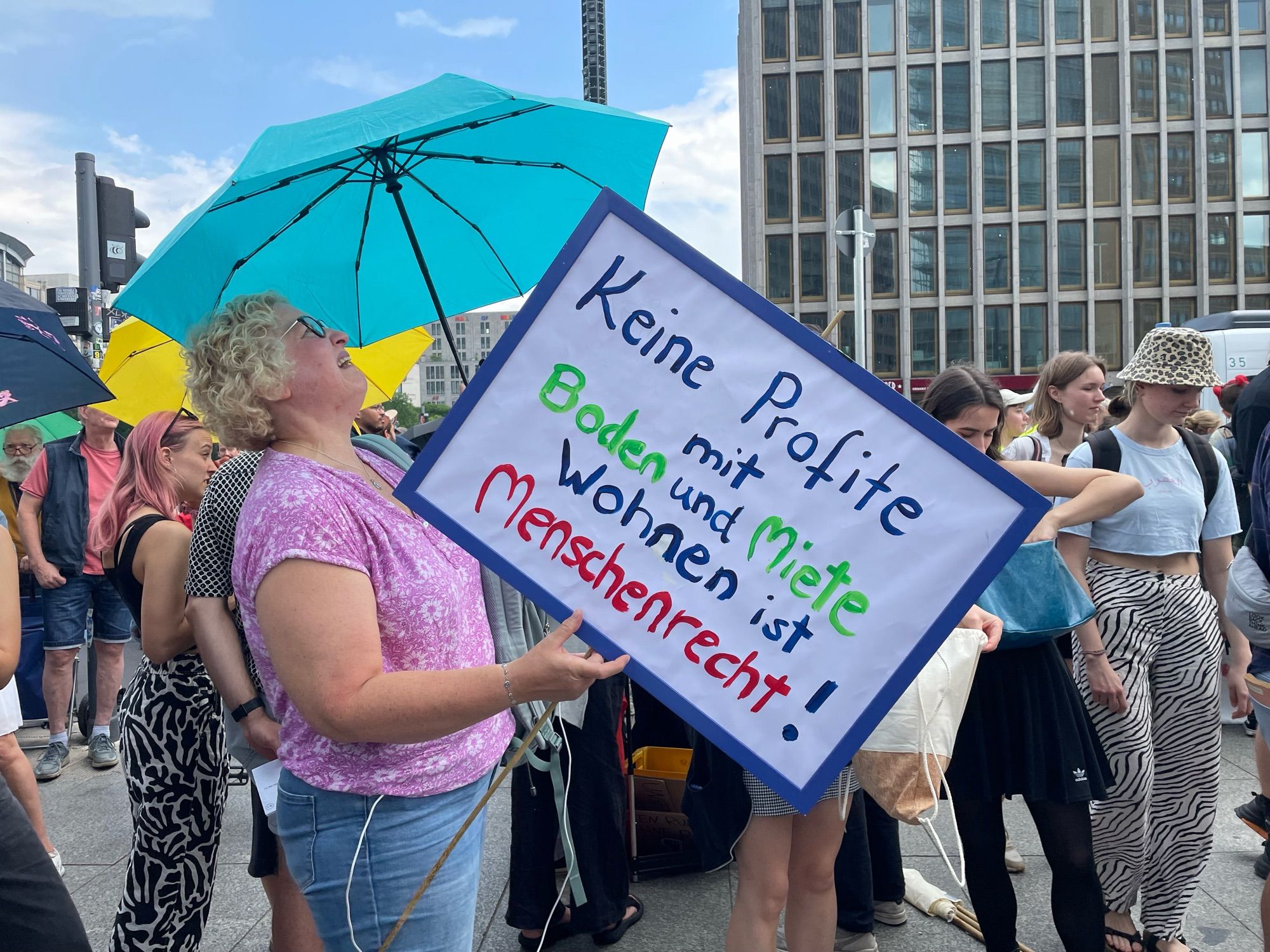 Eine blonde gelockte Frau blond vlt. unter einem blauen Regenschirm hoch und hält ein Plakat „Keine Profite mit Boden und Miete. Wohnen ist Menschenrecht!“