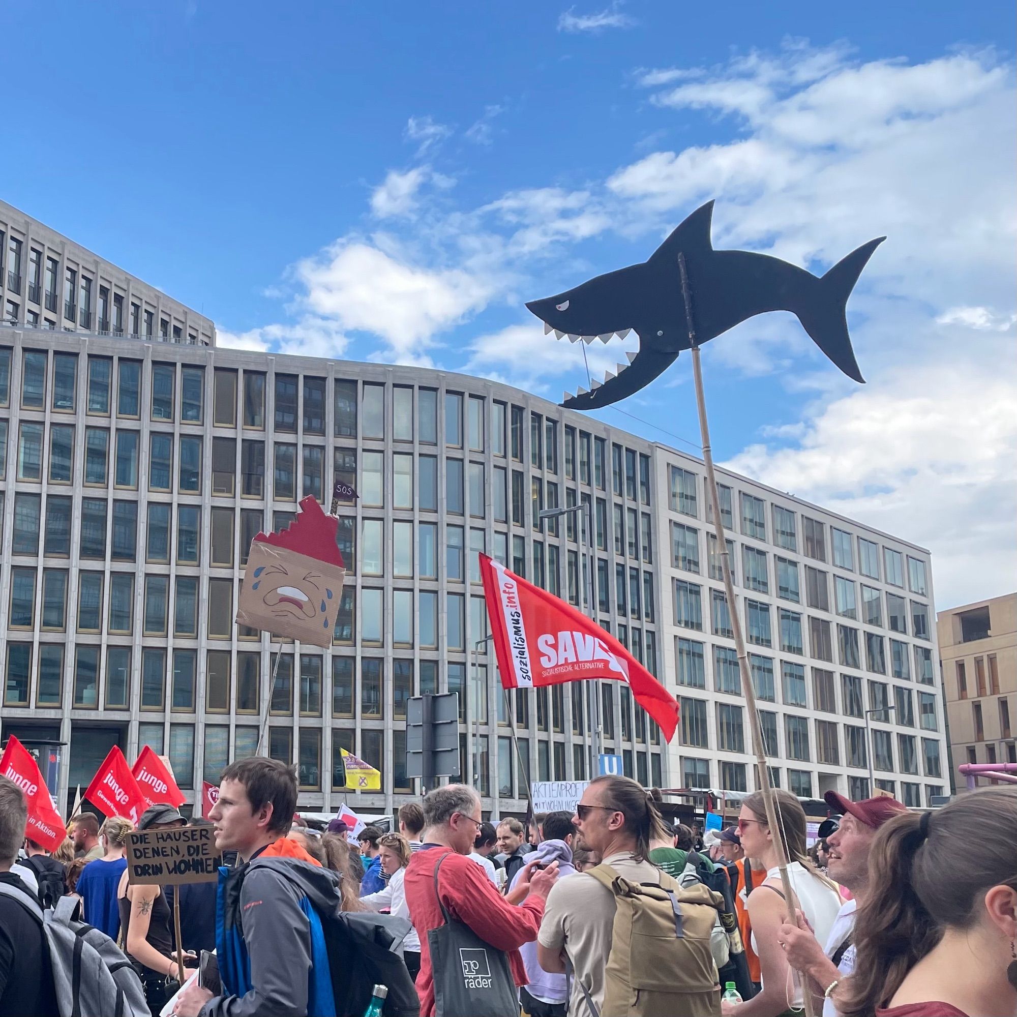 Demonstrierende vor einem Bürogebäude. In der Luft hängt ein schwarzer Hai mit offenem Maul und scharfen Zähnen.