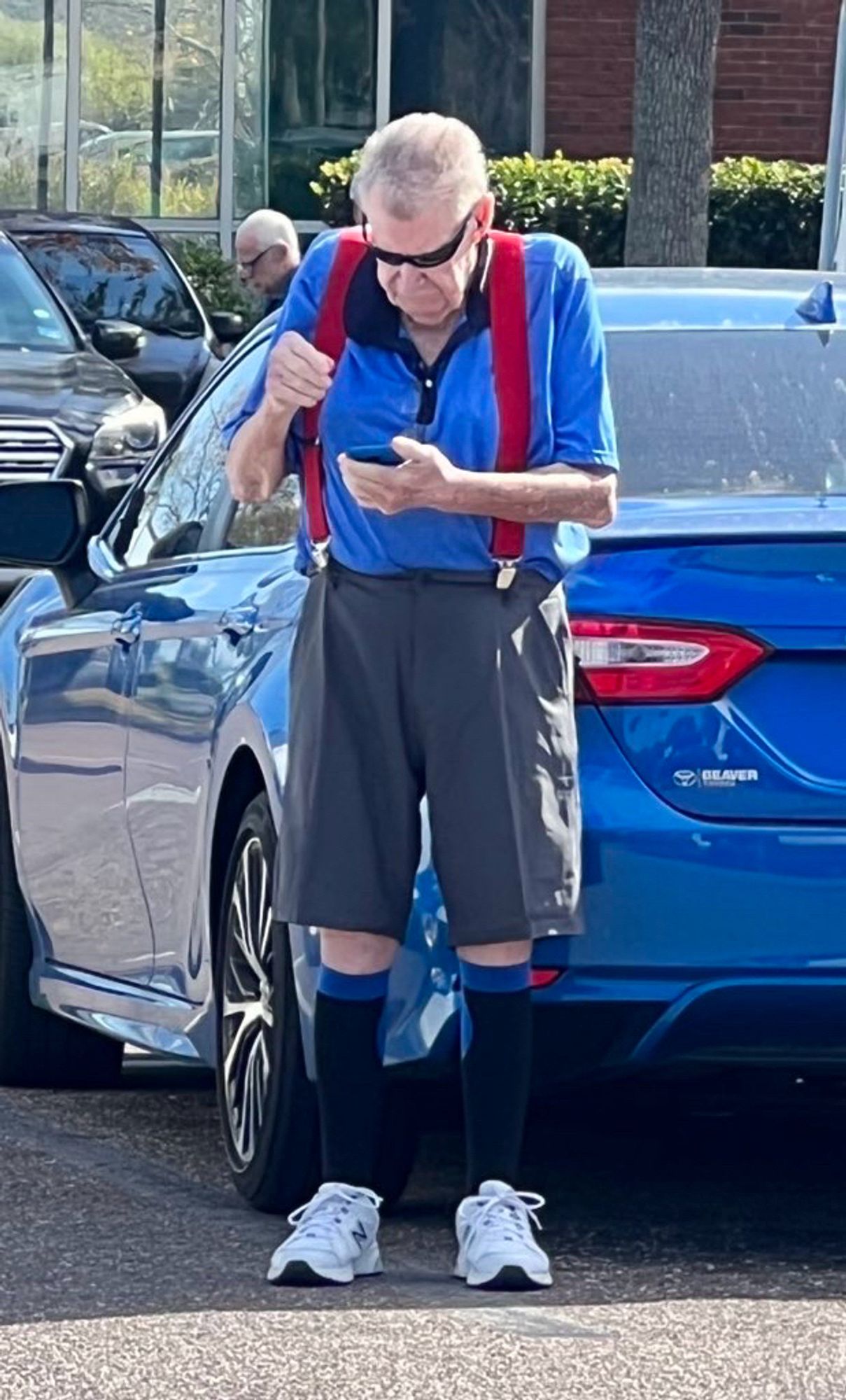 Older gentleman wearing athletic sneakers, knee high black socks with blue band, knee-length polyester trousers held up with red suspenders, a blue polo with black cuff, black sport sunglasses and looking sternly at his mobile phone.