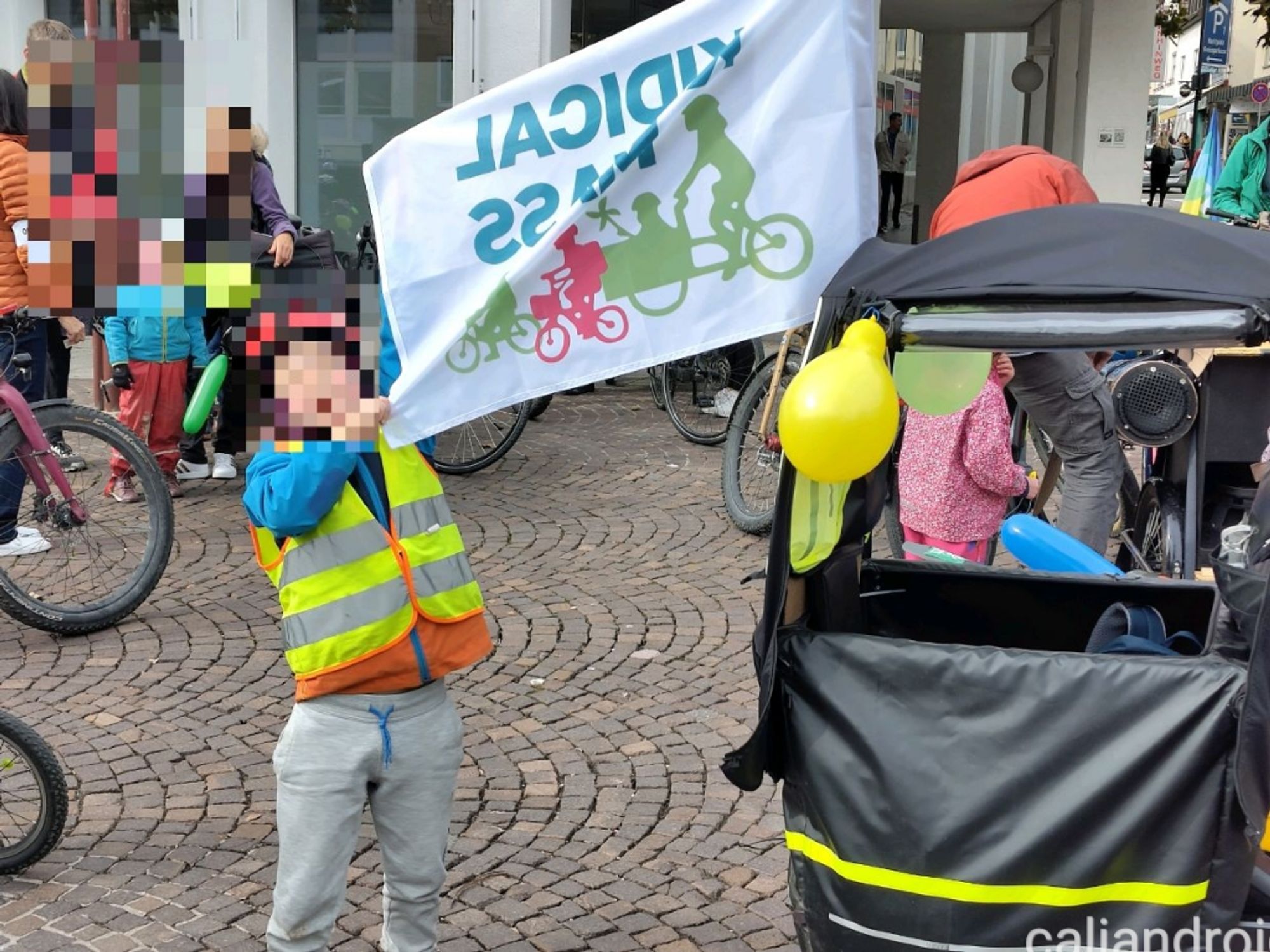 Sindelfinger Markplatz.
Ein Junge hebt eine KidicalMass Fahne hoch. Drumherum sammeln sich die Mitfahren den. An einem Longtail Lastenrad hängen bunte Ballons