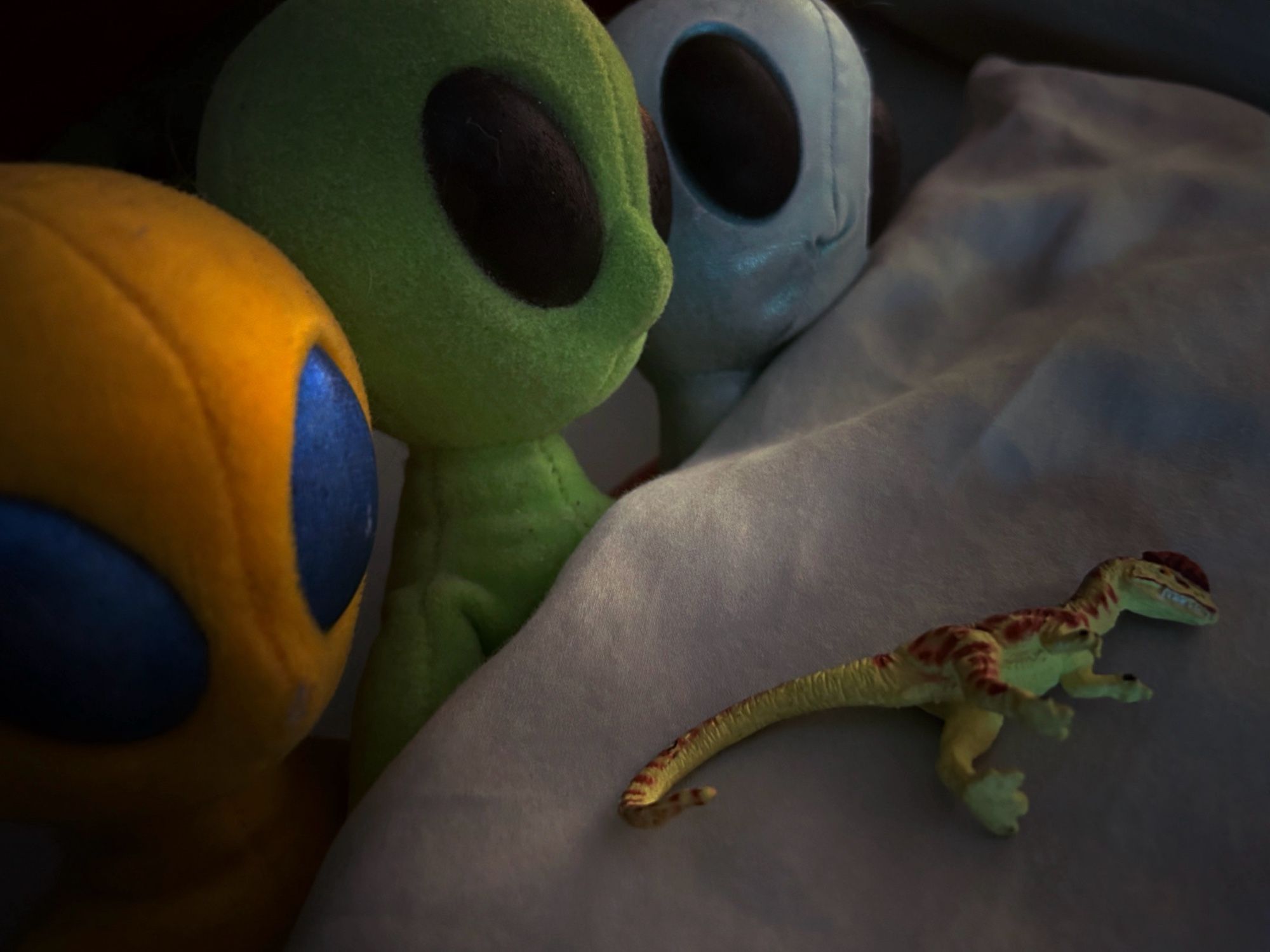 A green Dilophosaurus with brown dots lies sleeping on a light blue-green cushion while three extraterrestrials pop their heads out from the left side. The aliens from front to back are yellow, green, and blue with large almond eyes.