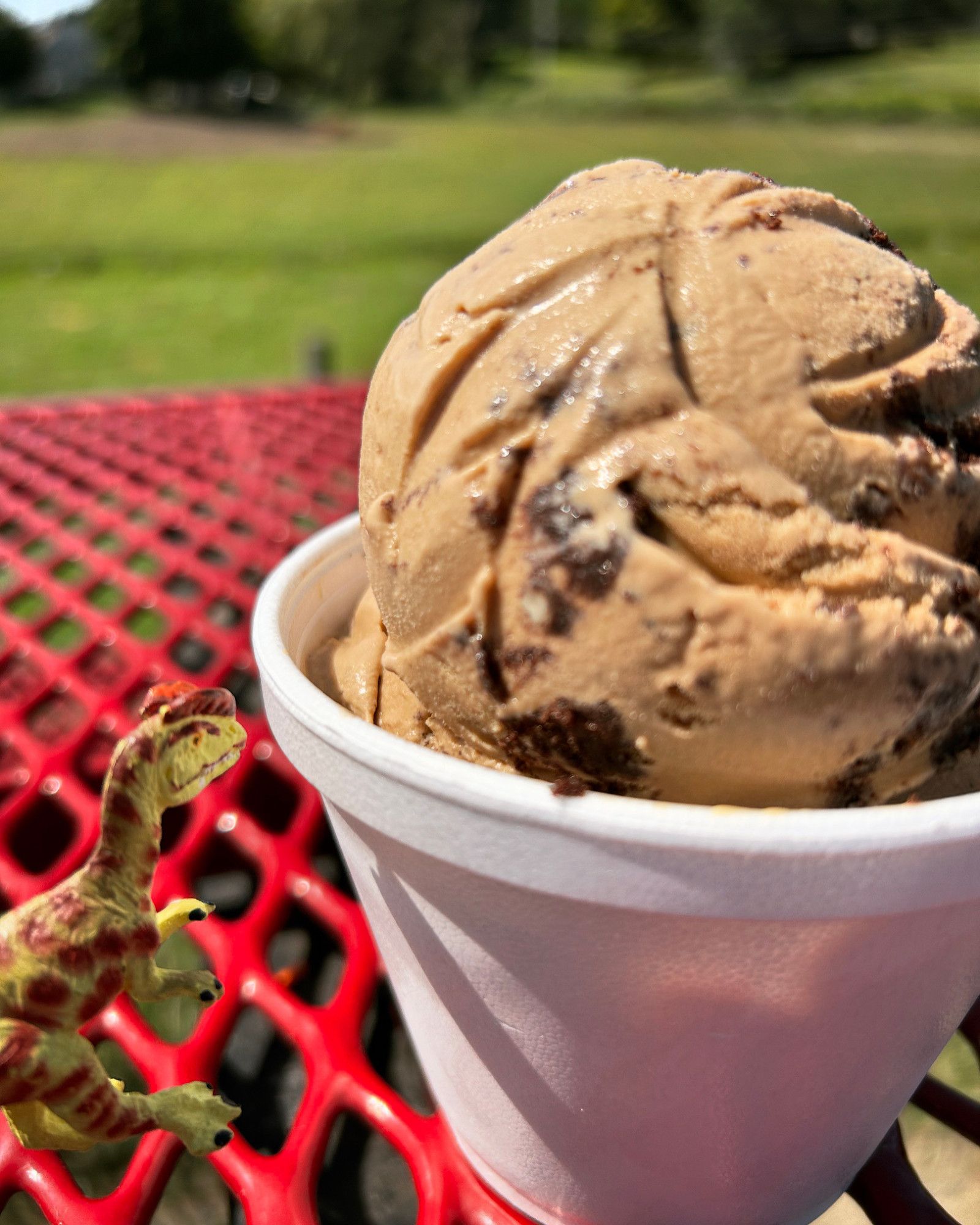 A green Dilophosaurus with brown spots eyeing a cup of coffee Oreo ice cream on a red picnic table. Green fields are behind them.