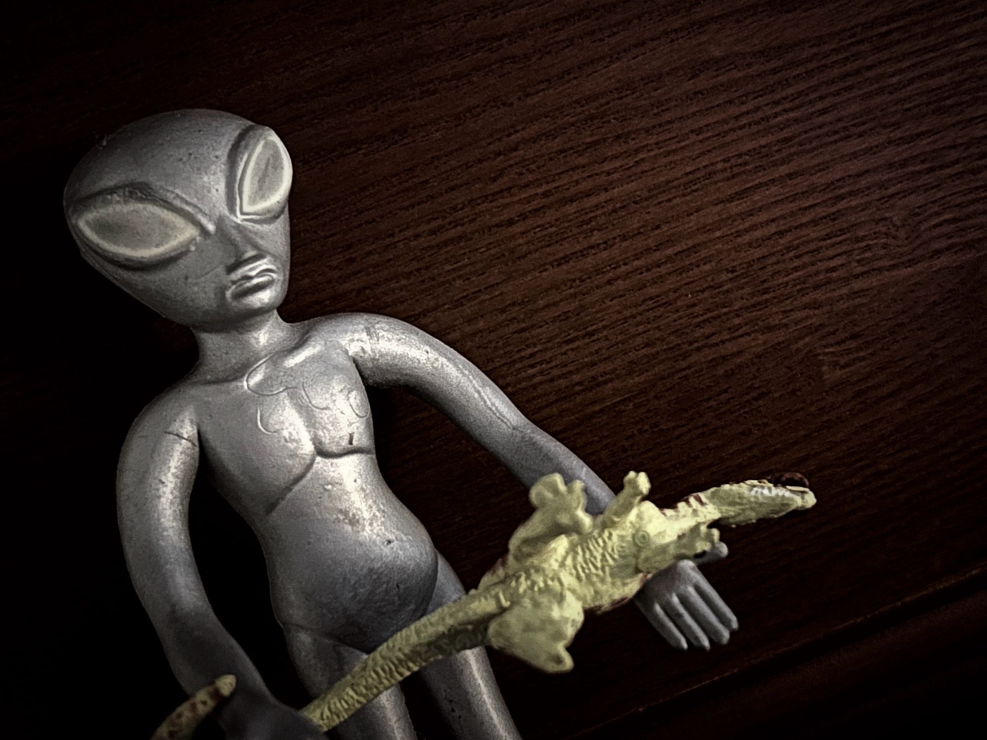 A grey alien from beyond the moon holding a stunned green Dilophosaurus in its arms.