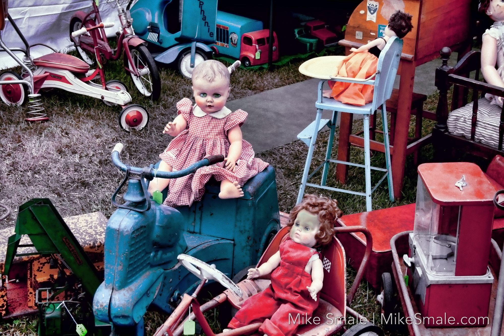 A creepy doll with lime green sitting atop a derelict toy scooter. Various junk surrounds her including another doll with burns on her face.