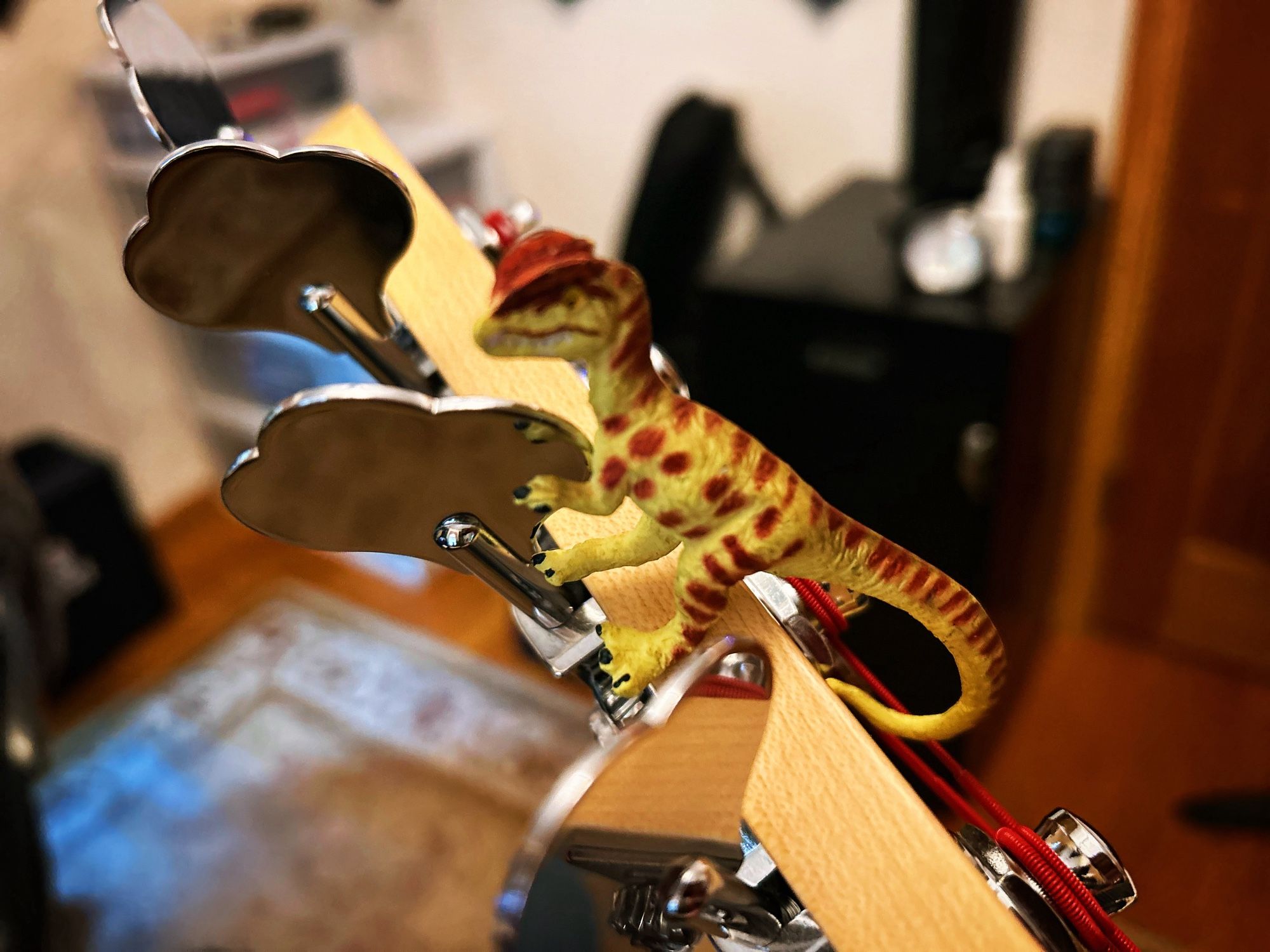 A green Dilophosaurus with brown spots holding onto the tuning machine of a Fender bass guitar with red strings. A file cabinet, air purifier, and bottle of fingerboard oil can barely be seen in the background.