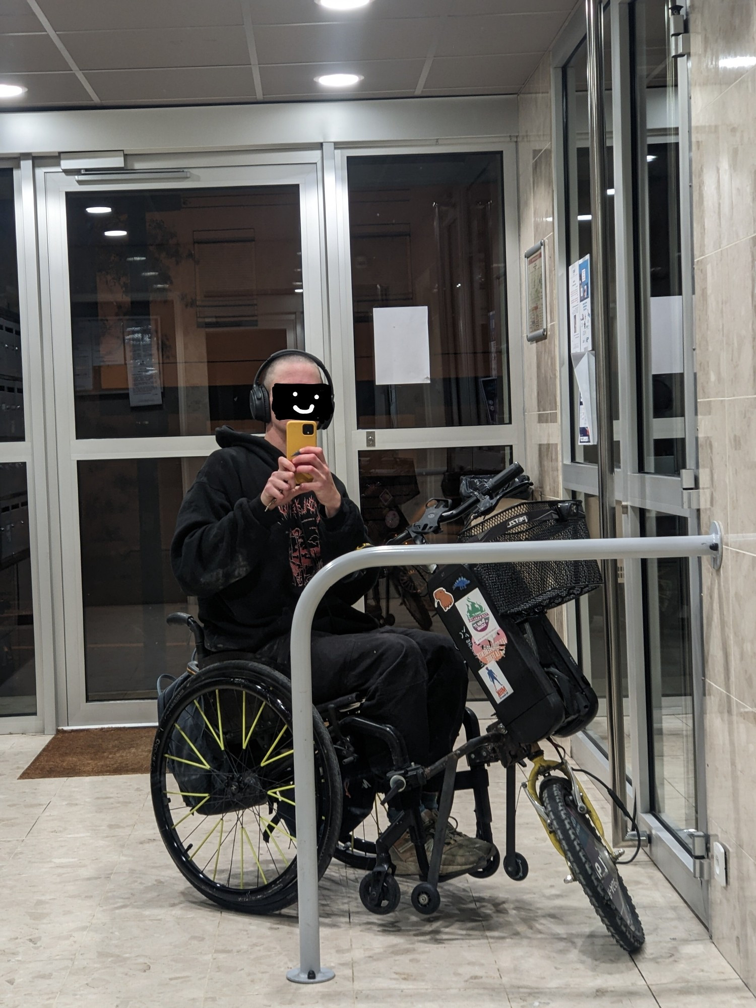 Selfie miroir dans un hall d'immeuble, je suis habillé en noir assis sur mon fauteuil roulant actif noir avec des rayons jaunes, devant il y a un système de motorisation avant avec une grosse roue, deux batteries de vélo électrique et un guidon, il y a des autocollants sur les batteries