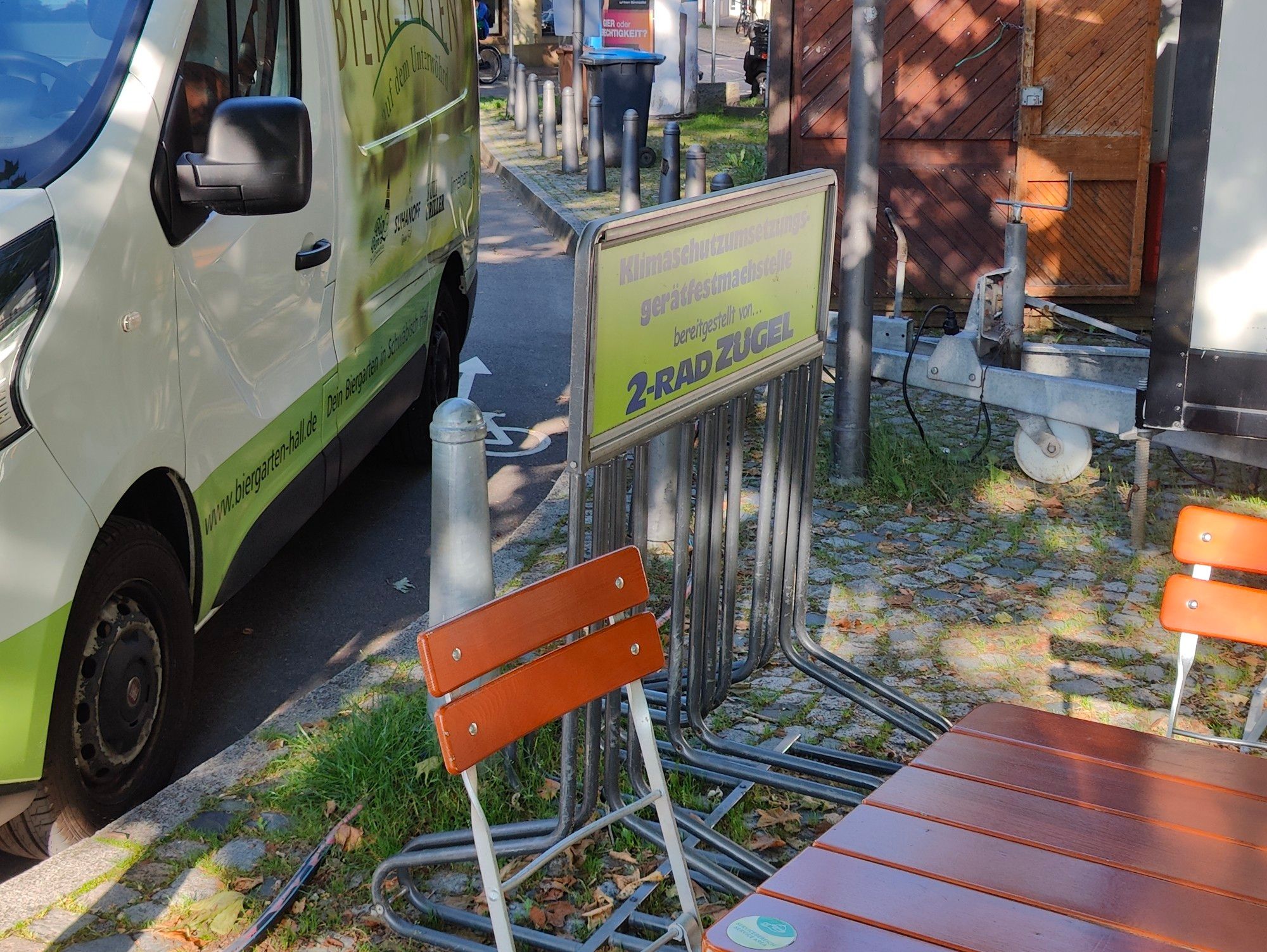 Foto auf dem ein Auto auf dem Fahradwegzeichen parkt neben einem Fahrradabstellgatter, das so zwischen Poller und Biergartentischen eingeszwängt ist, dass man dort unmöglich ein Fahrrad abstellen kann