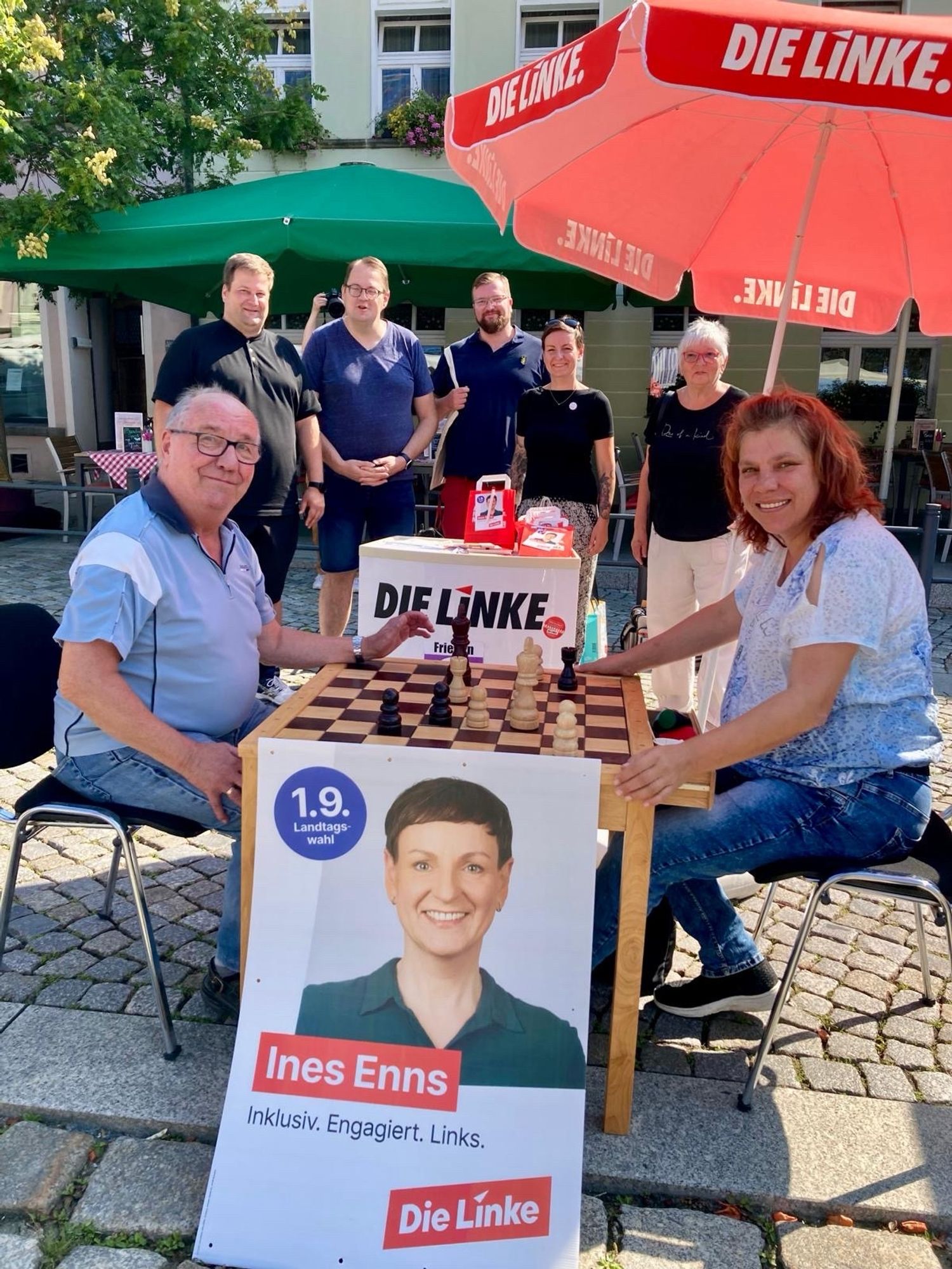 Im Vordergrund, zwei Personen an einem Schachttisch. Im Hintergrund ein Sonnenschirm DIE LINKE und fünf weitere Personen.