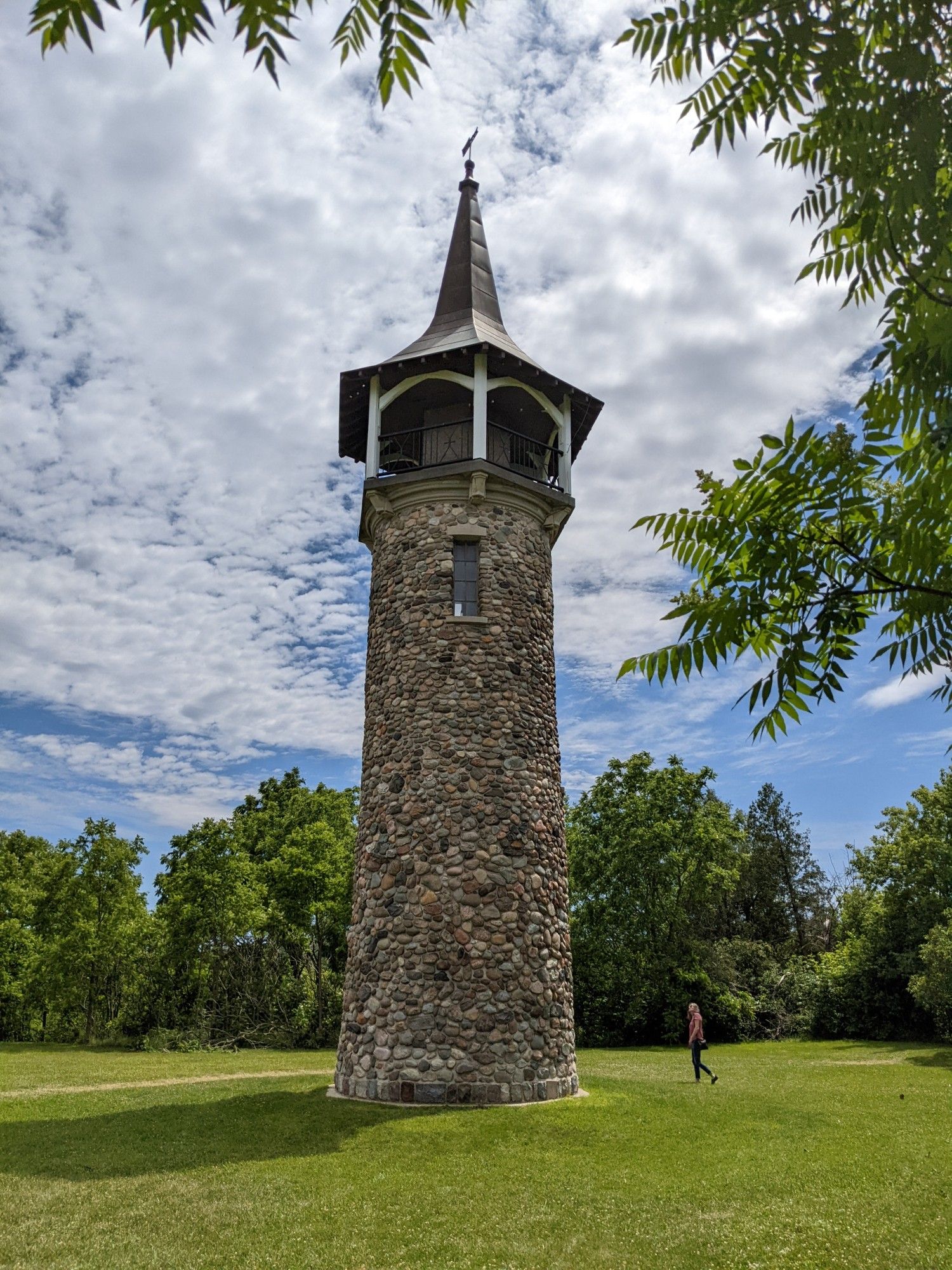 A tall cylindrical stone tower, with an open viewing area at the top, under a pointed, conical metal roof that culminates in a weather vane. The walls are made of rounded field stones mortared together. There is one tall, thin, window visible high up the tower, and no visible entry. Overall the effect is of a tower wearing a wizard hat.

There is a large, roughly circular, sward of lawn around the tower, which is in turn surrounded by trees.  A person is shown near the tower, establishing scale: the tower is 5-6 times the height of a person, not including the roof.