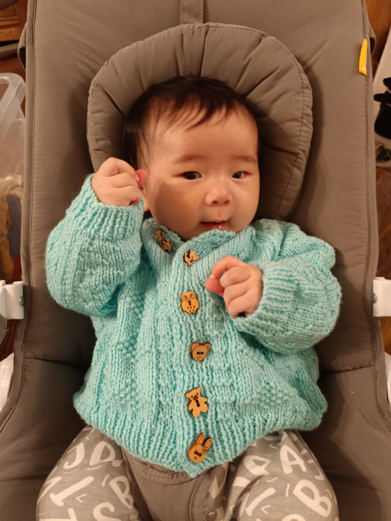 Baby wearing a turquoise coloured, knitted cardigan with wooden animal buttons, sat in a rocker with right fist up and looking coyly at the camera