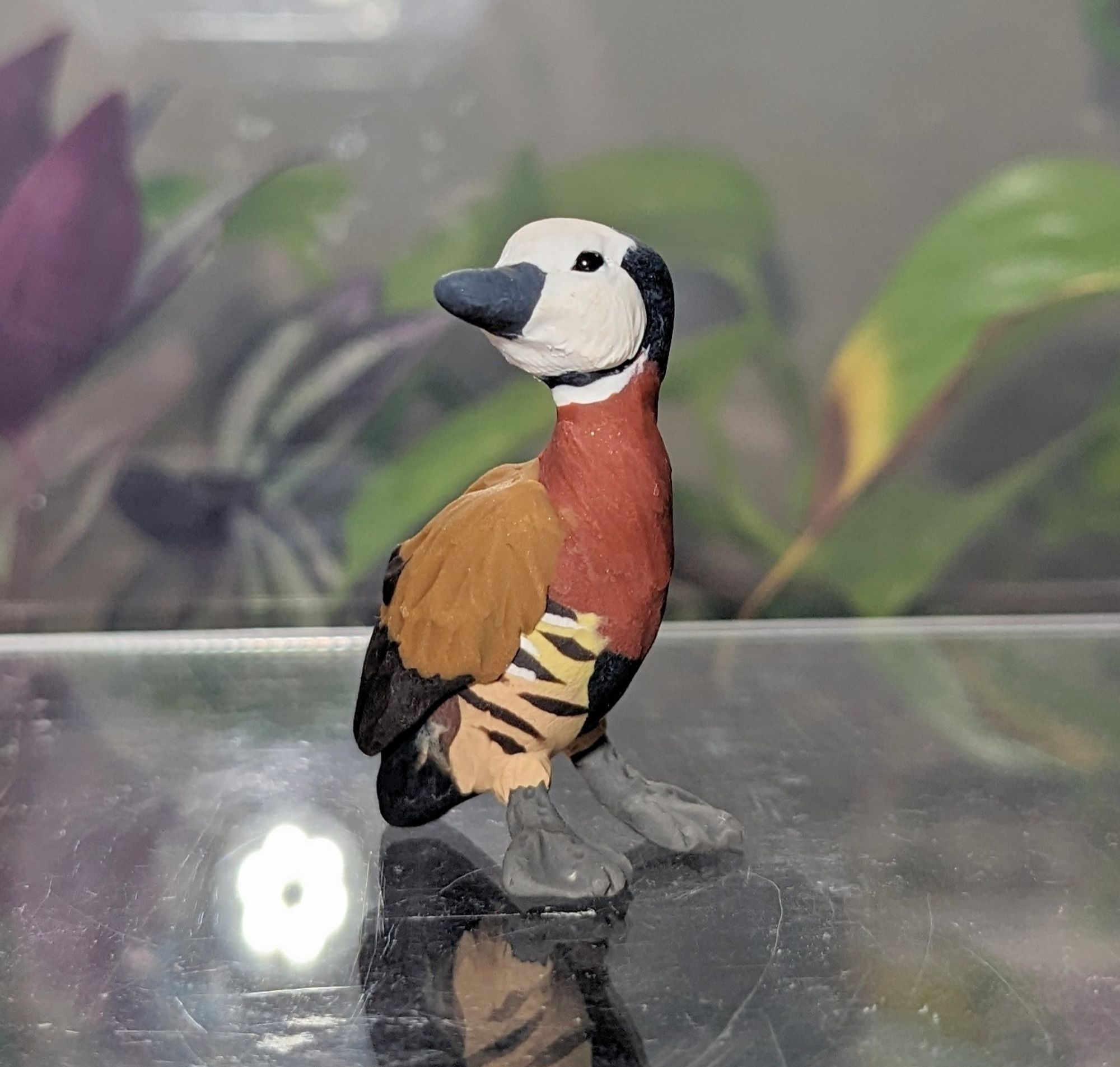 A figurine of a colourful duck, head turned upward and to the side, to better watch the viewer. The duck has a white face a gray bill, a reddish brown chest, brown wings, and a striped underside. The duck also has a black chin strap, and looks alert and lively.