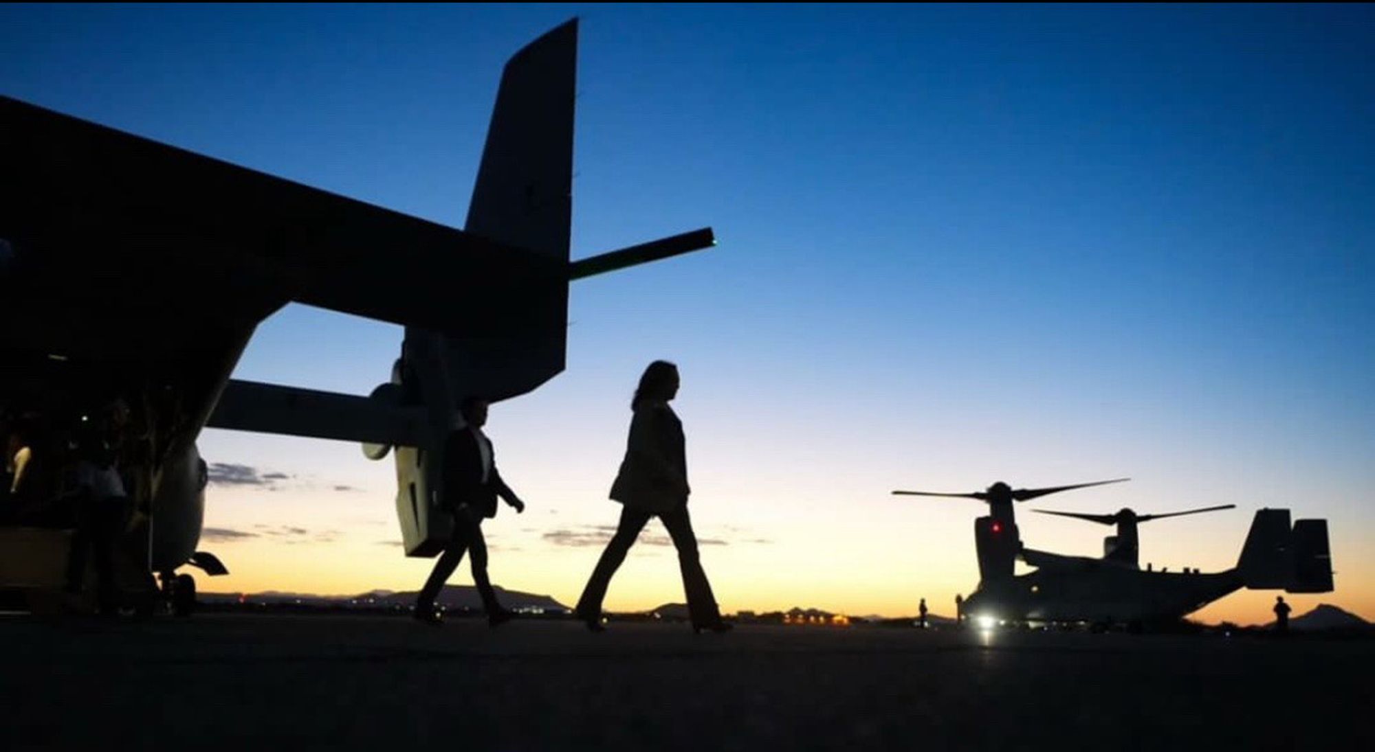 Silhouettes of people walking on a tarmac at sunset with military aircraft in the background.