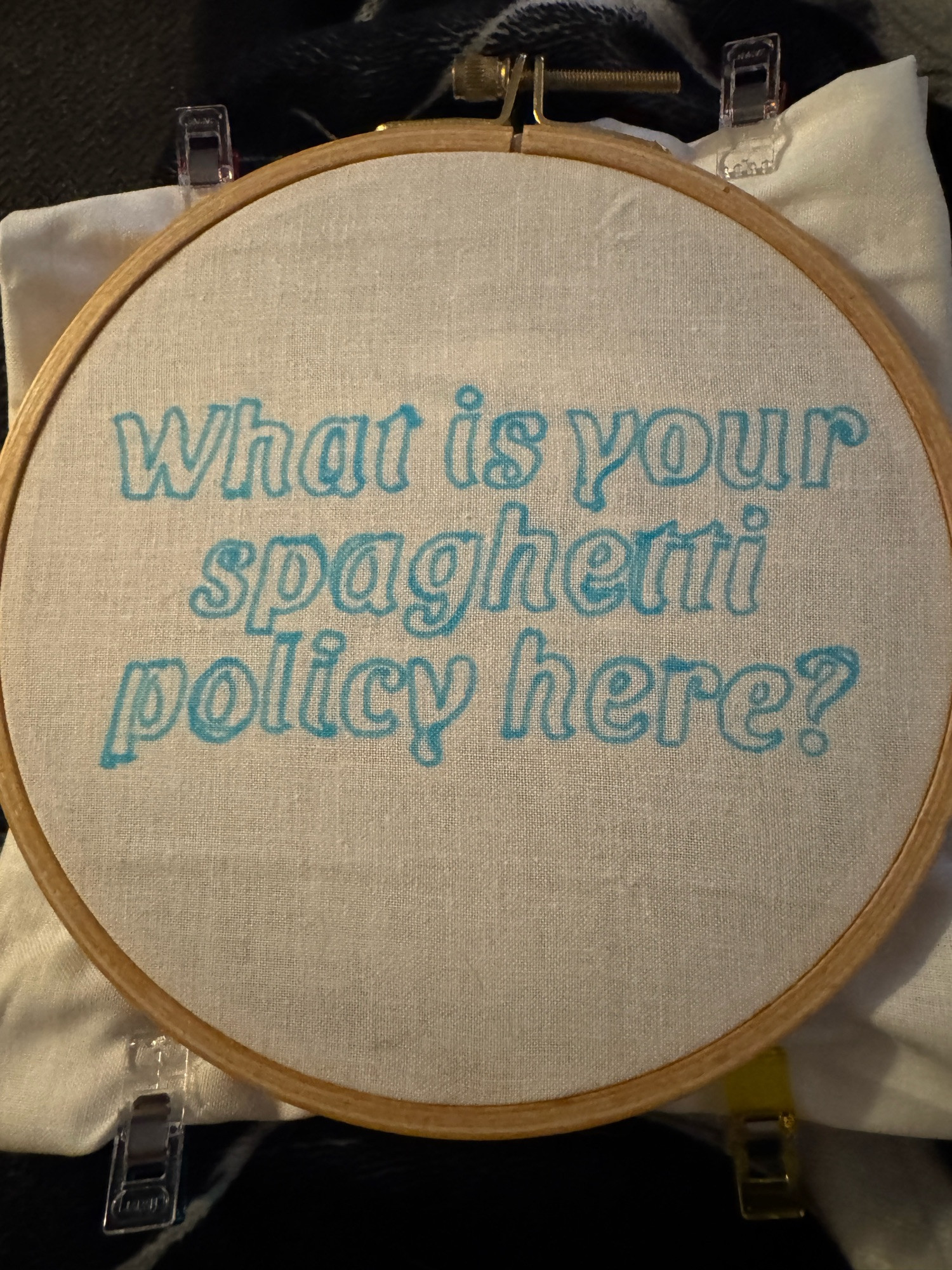 The outline of text on fabric that reads, “What is your spaghetti policy here?” The fabric is on an embroidery hoop, ready to be filled in