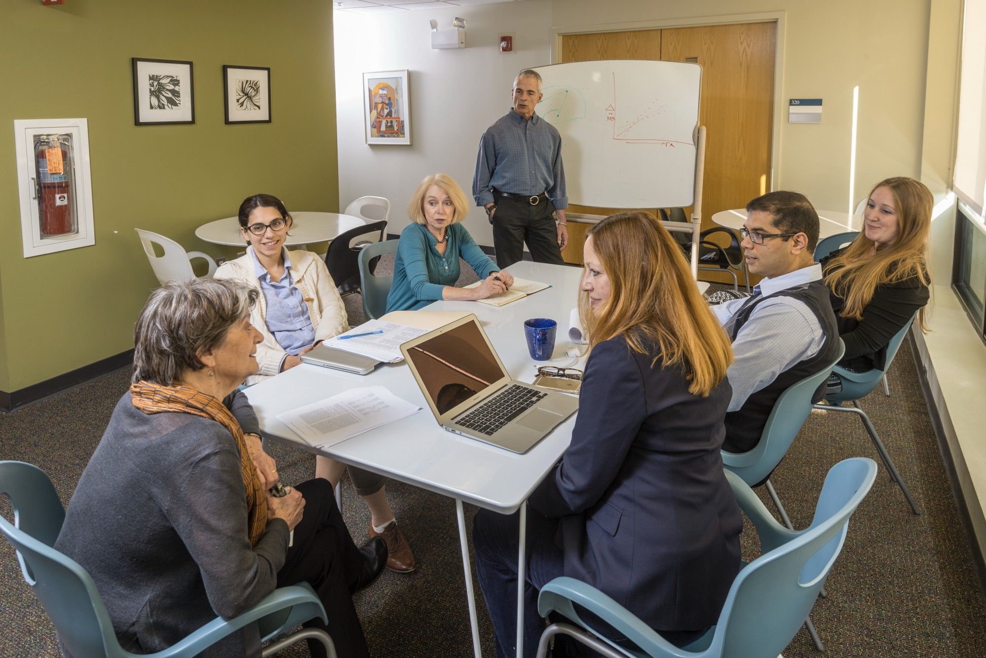 This image shows seven MRRI scientists seated at a conference room table. One scientist is standing in the back in the middle in front of a white board, and the six seated scientists are engaged in discussion. 