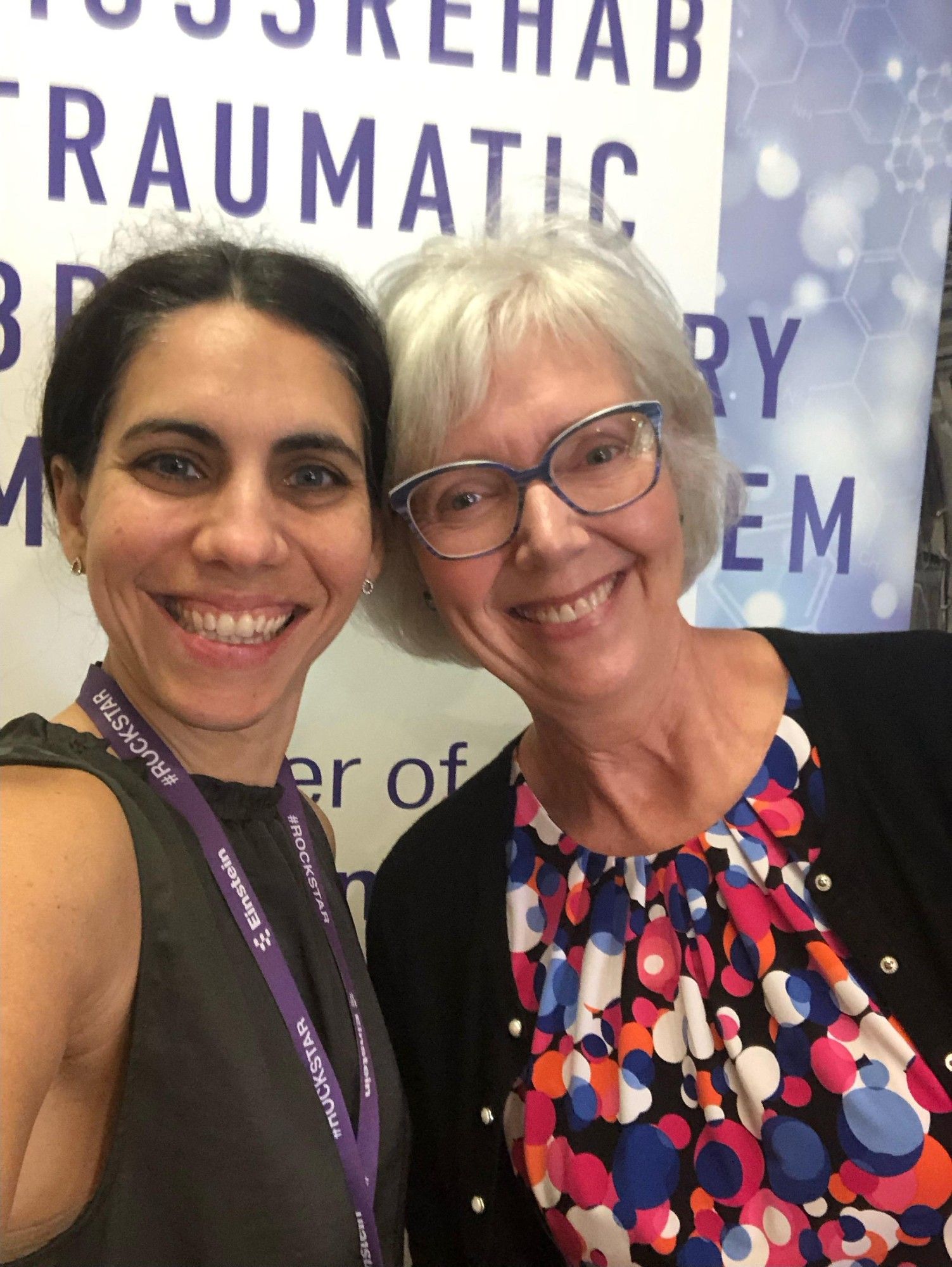 This photo features Dr. Amanda Rabinowitz (left) and Dr. Lyn Turkstra (right). Both are smiling and looking at the camera. There is a banner behind them for the MossRehab Traumatic Brain Injury Model System. 