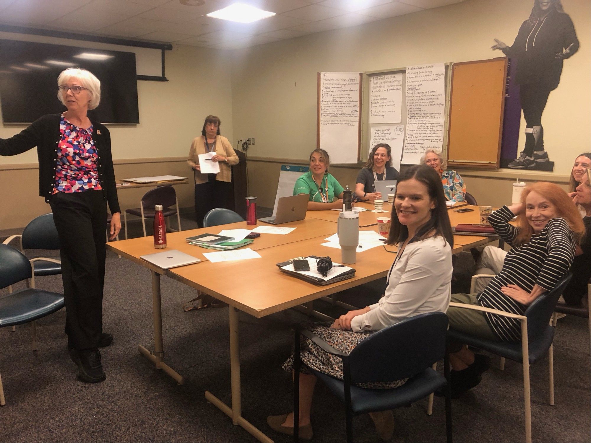 Dr. Lyn Turkstra is presenting to a group of team members working on this project. Dr. Turkstra is standing on the left pointing at something out of frame. One team member is standing in the back of the image, and the remaining team members are seated at a conference table. 