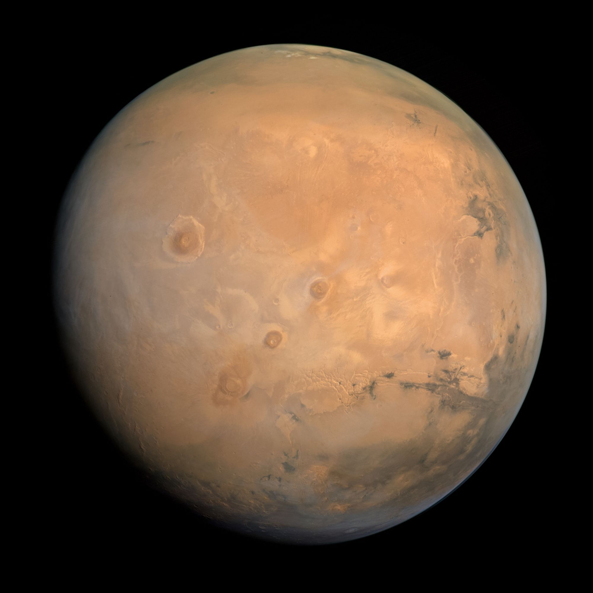 Mars as imaged by the Hope orbiter. The planet is mottled rusty orange. The massive shield volcanoes are visible near the equator