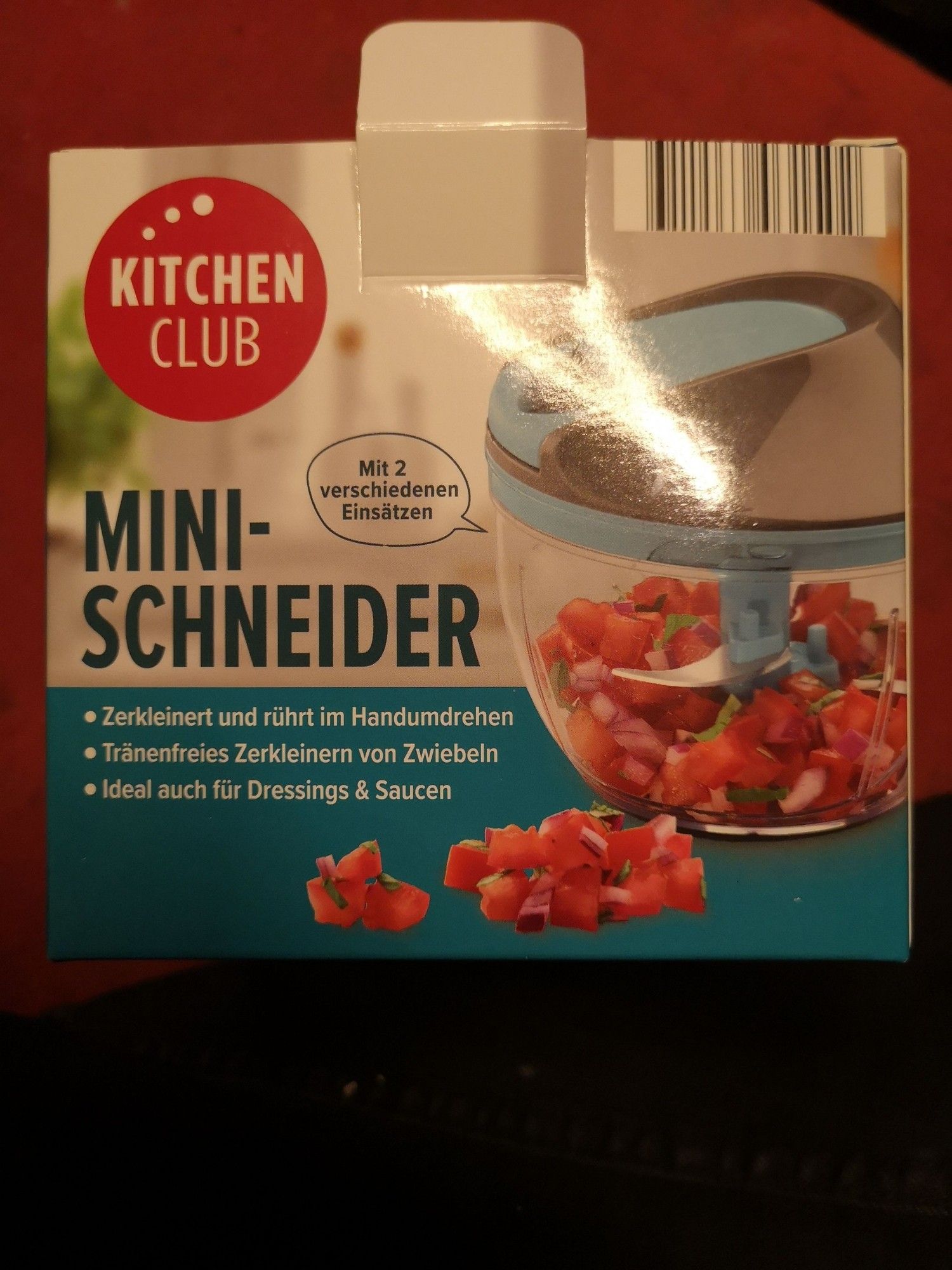 Die Packung des Mini-Schneiders
Im unteren Drittel türkis, oberhalb grau. Seitlich ist das Gerät abgebildet: zu erkennen eine durchsichtige Schale mit türkisgrauem Deckel, darin sind die Messer, zerkleinerte Tomaten und Kräuter zu sehen. 
Als Text steht drauf:
Kitchen Club
Mini-Schneider
Zerkleinert und rührt im Handumdrehen
Tränenfreies Zerkleinern von Zwiebeln
Ideal auch für Dressings und Saucen
Neben dem Gerät eine Sprechblase in der steht: mit 2 verschiedenen Einsätzen