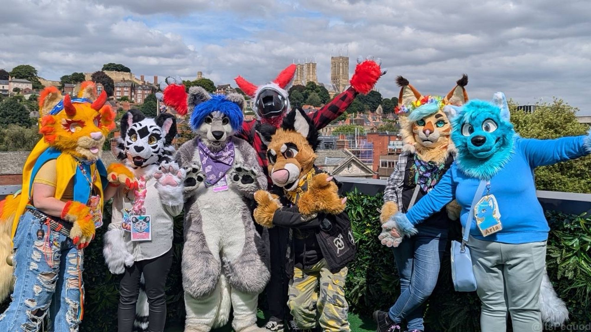 A group of fursuiters posing on a green balcony decorated with leaves. The scene behind is a cloudy, summer sky with Lincoln Cathedral in the background.