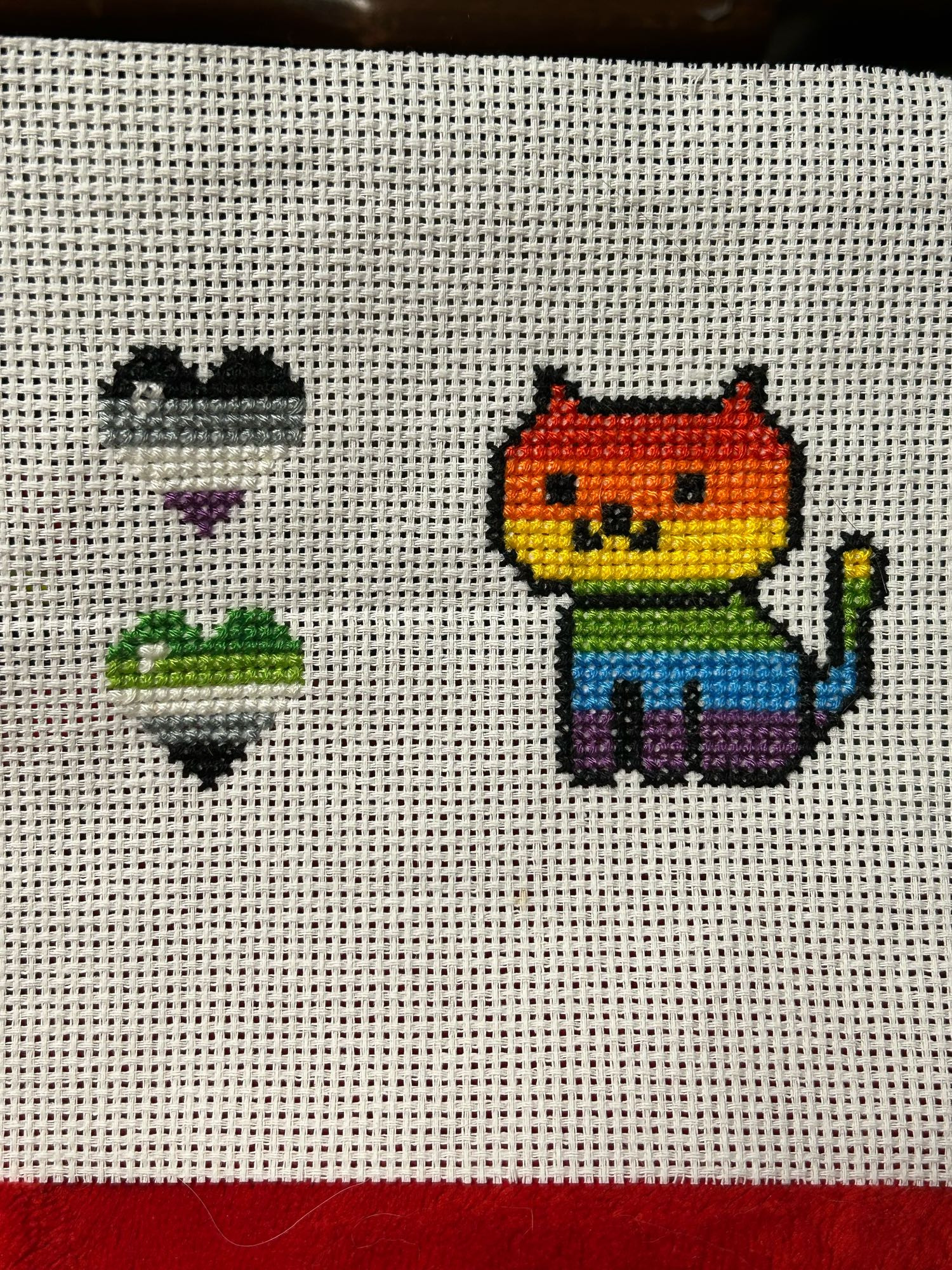 White Cross stitch canvas with two small cross stitched hearts on the left and a cross stitched cat on the right. The hearts are made with the Ace flag colours (top) and Aro flag colours (bottom); the cat is filled in with the rainbow. All colours go horizontal