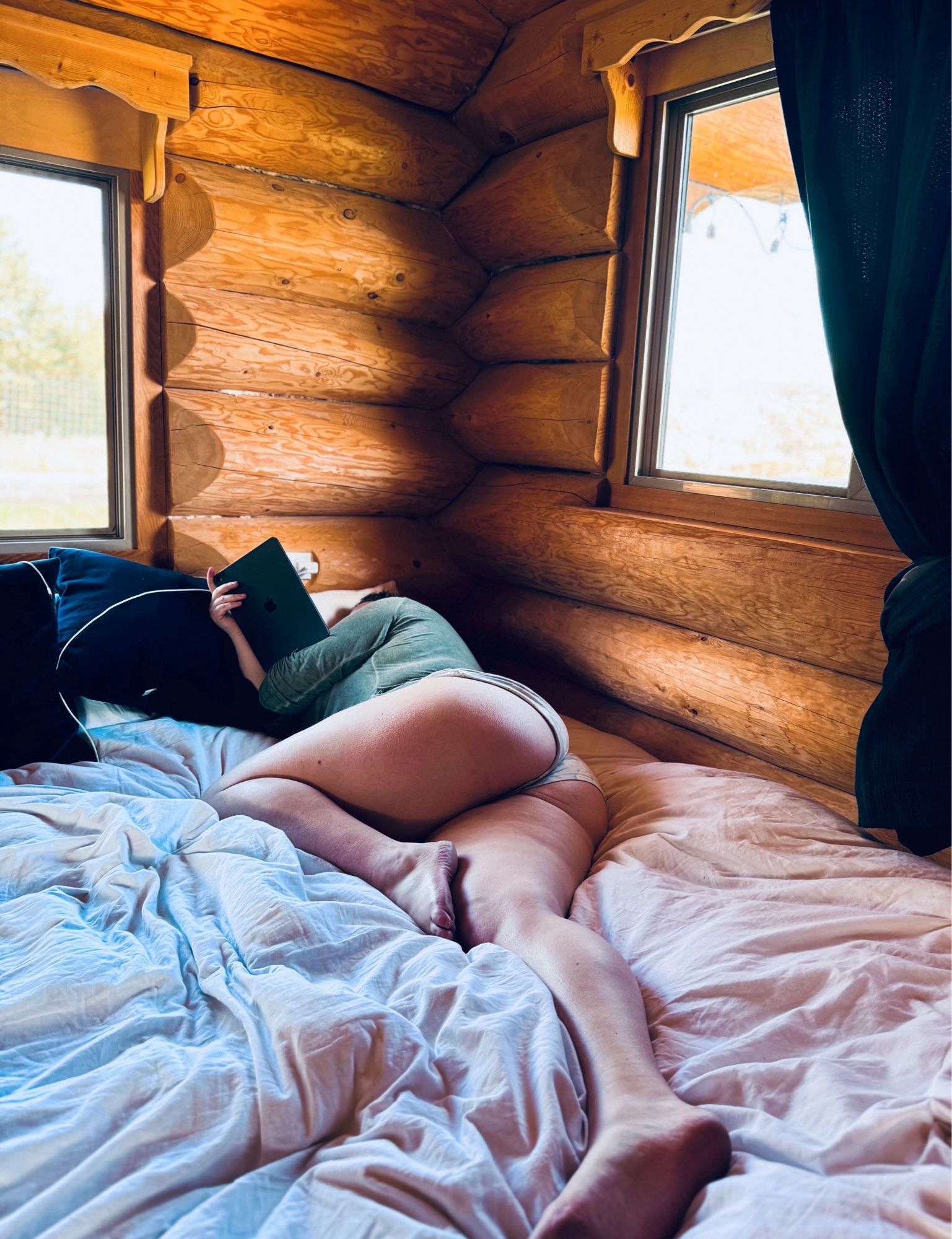 A shot of me lying in bed having a cozy lazy morning reading. I’m I’m a big bed with white sheets in a cozy log cabin with big windows. I’m wearing pj shorts and a green t-shirt. My face is hidden but my booty and long legs are on display.