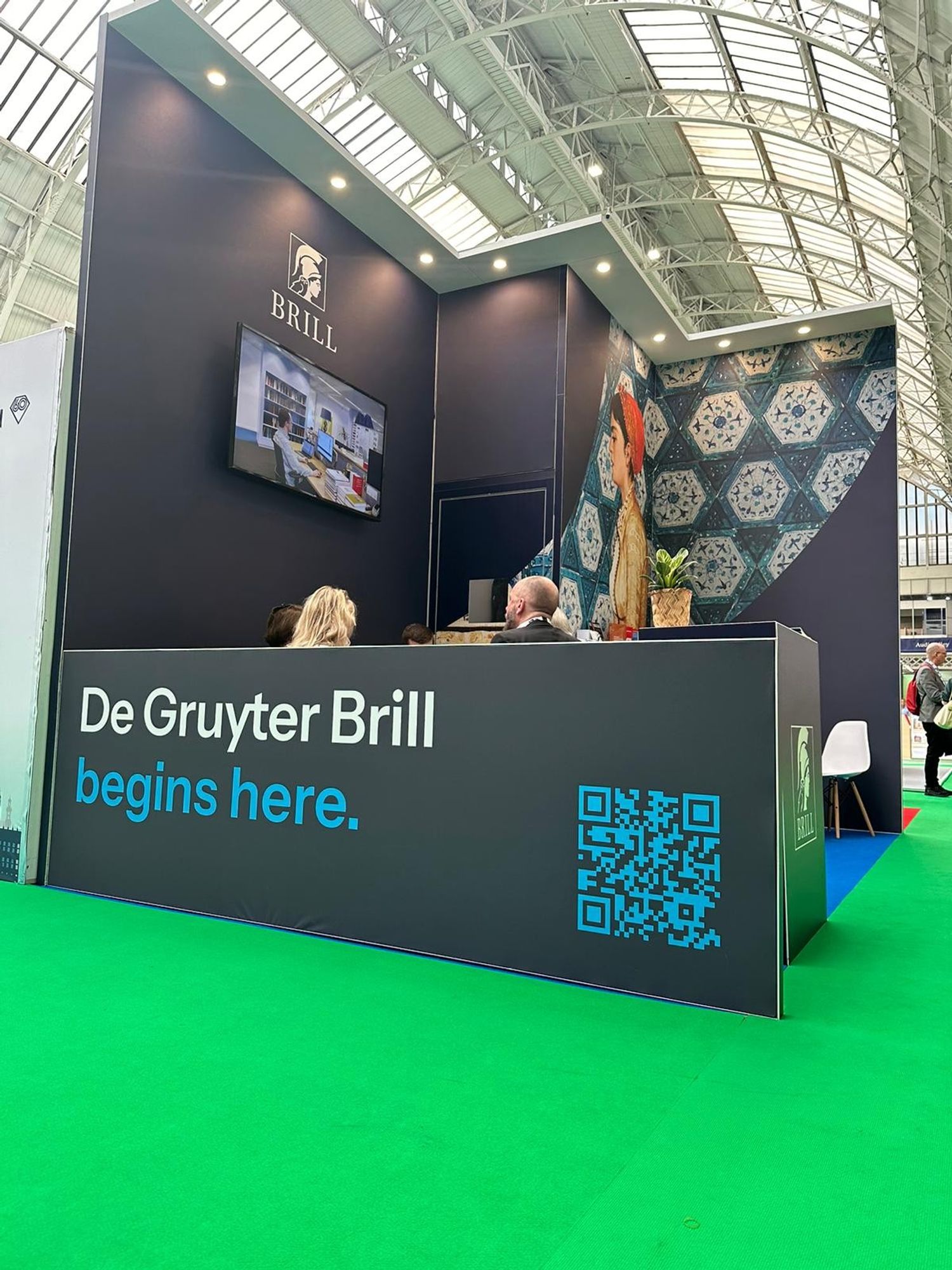 The Brill stand at London Book Fair. The Brill logo is seen above a TV screen, below which colleagues are seated in discussion. On the outside of the stand it reads, De Gruyter Brill begins here, next to a QR code.