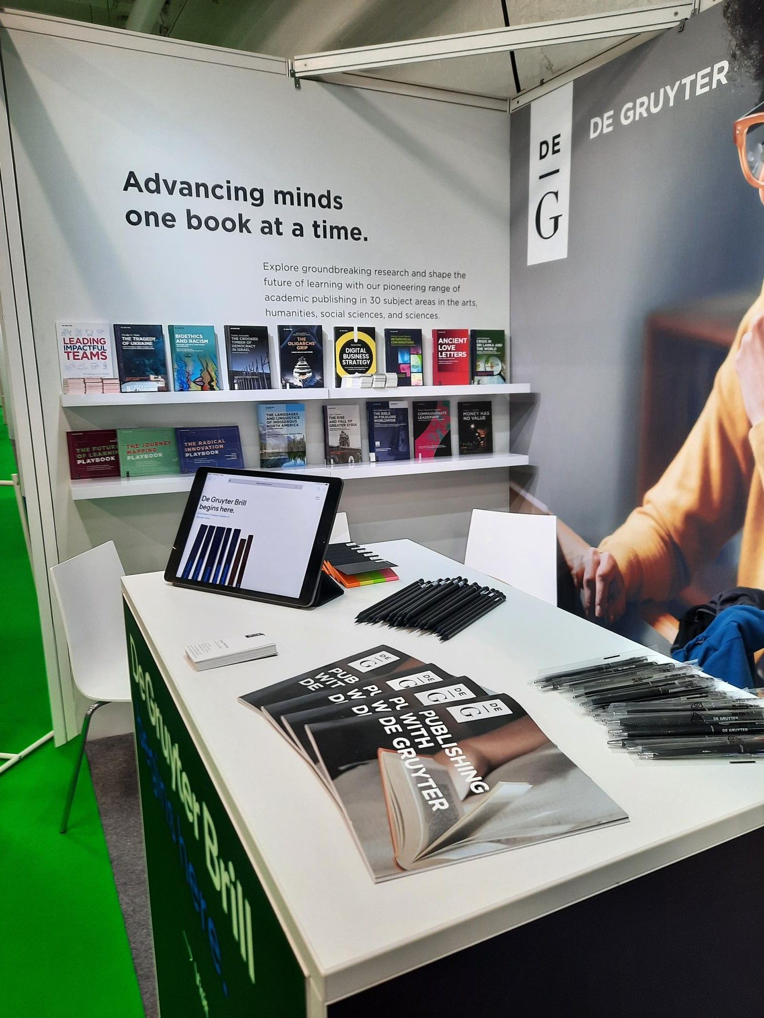 De Gruyter merchandise is seen on a desk at the De Gruyter stand. In the background, we see the De Gruyter logo next to two shelves of books. The display also reads, "Advancing minds one book at a time."