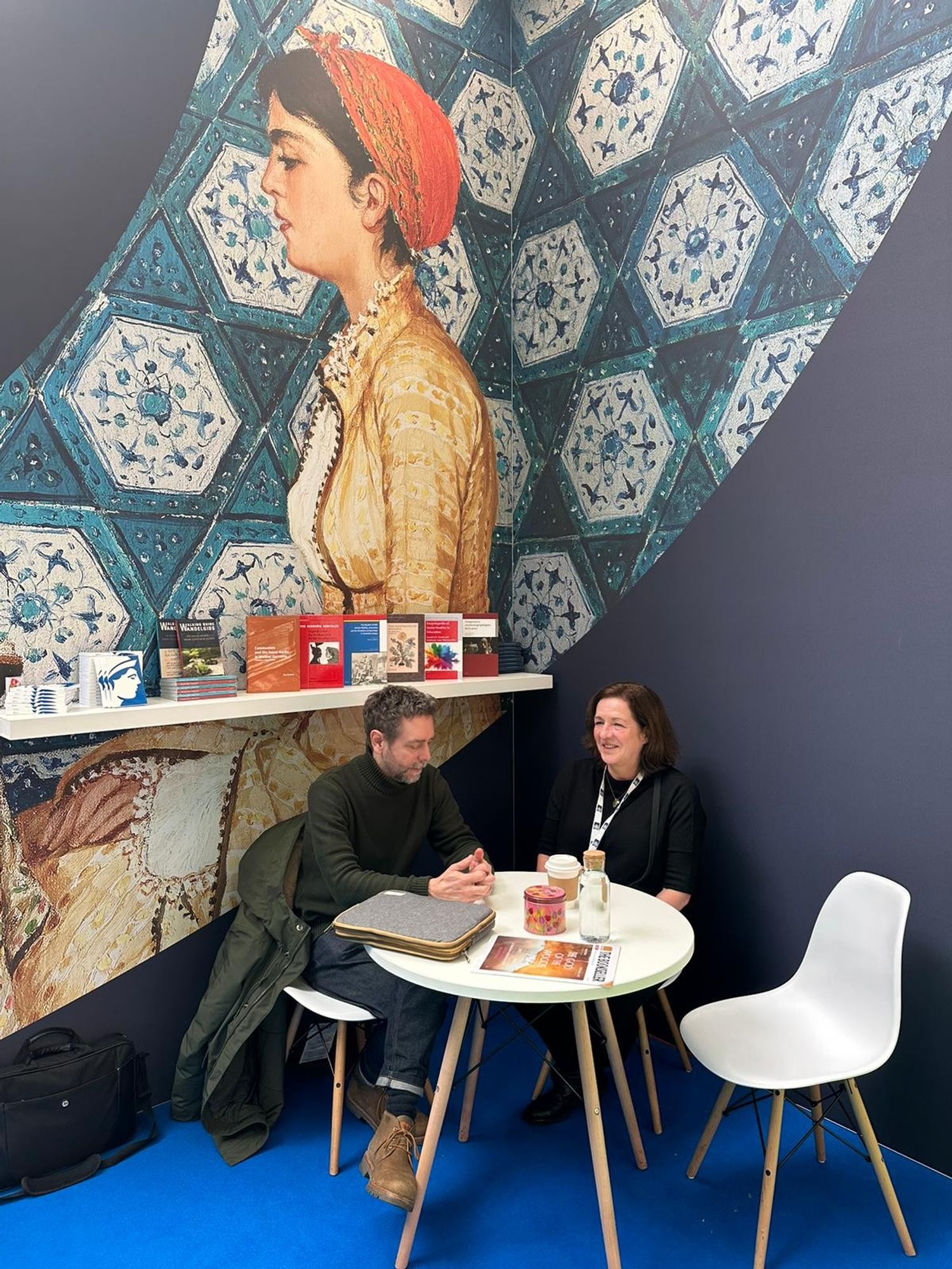 Two colleagues are seated in discussion at the Brill Stand, beneath a selection of books. In the background is a large illustration of a women in traditional dress against a tiled wall.