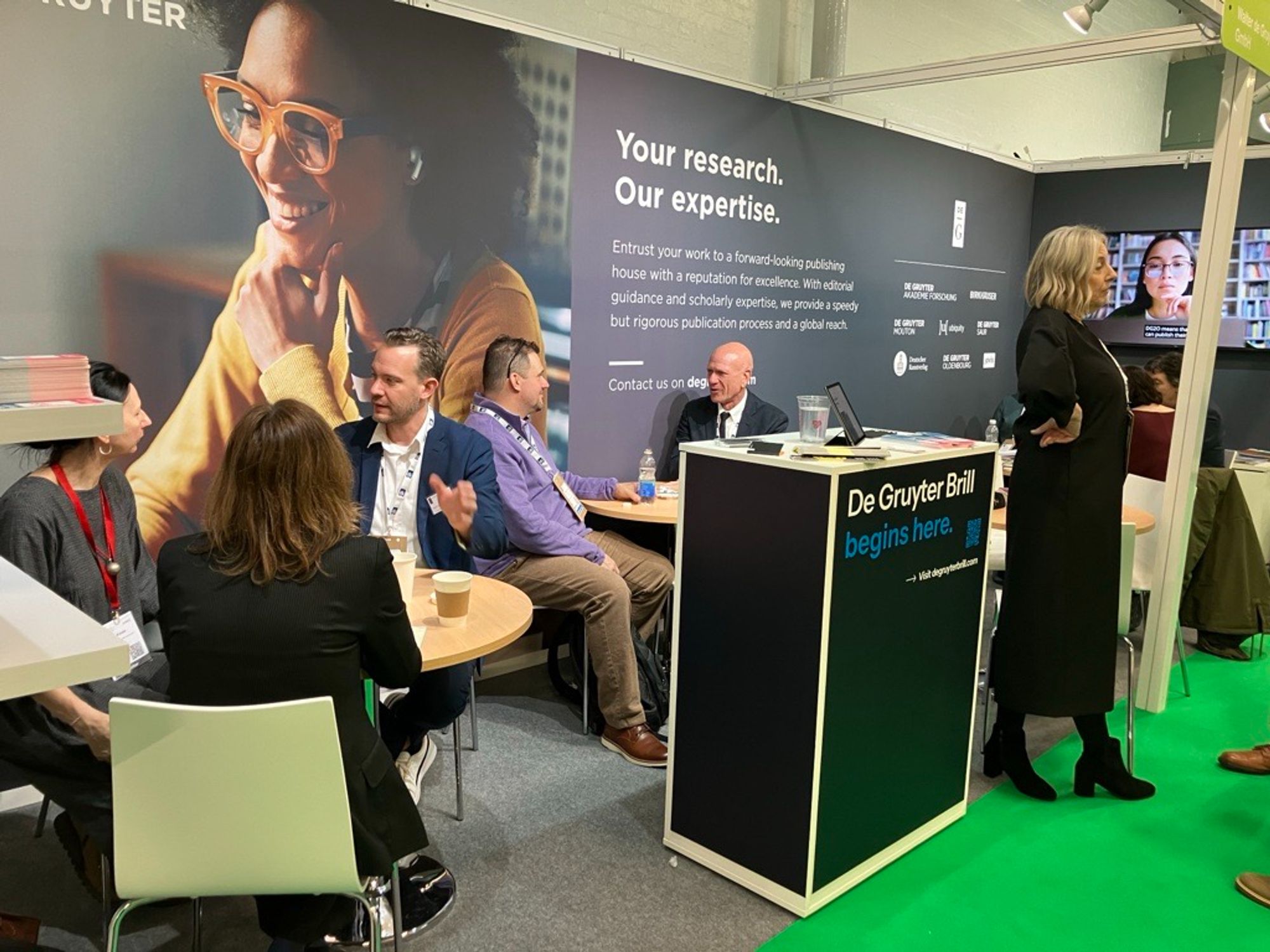 Colleagues are seated in discussion at the De Gruyter stand at the London Book Fair. A small desk display reads "De Gruyter Brill begins here." In the background, a large visual shows a smiling scholar next to the words, "Your research. Our expertise."