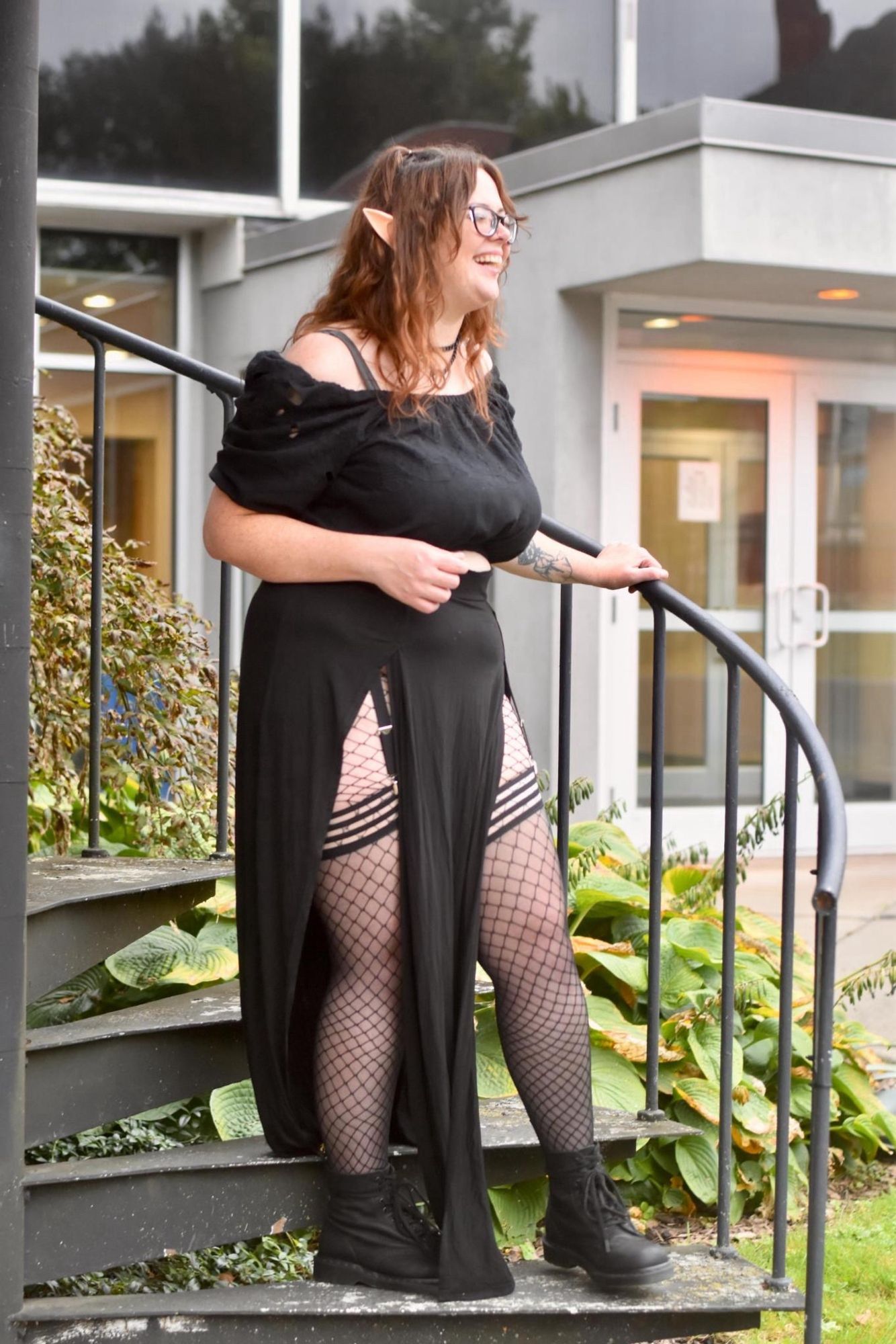 iris is pictured standing on a spiral staircase outside a museum, laughing in a candid capture. they're wearing all black and elf ears. they have combat boots and fishnets on, showing their full leg up to the waist with a long black skirt. they're wearing a garter belt and black martingale collar also.